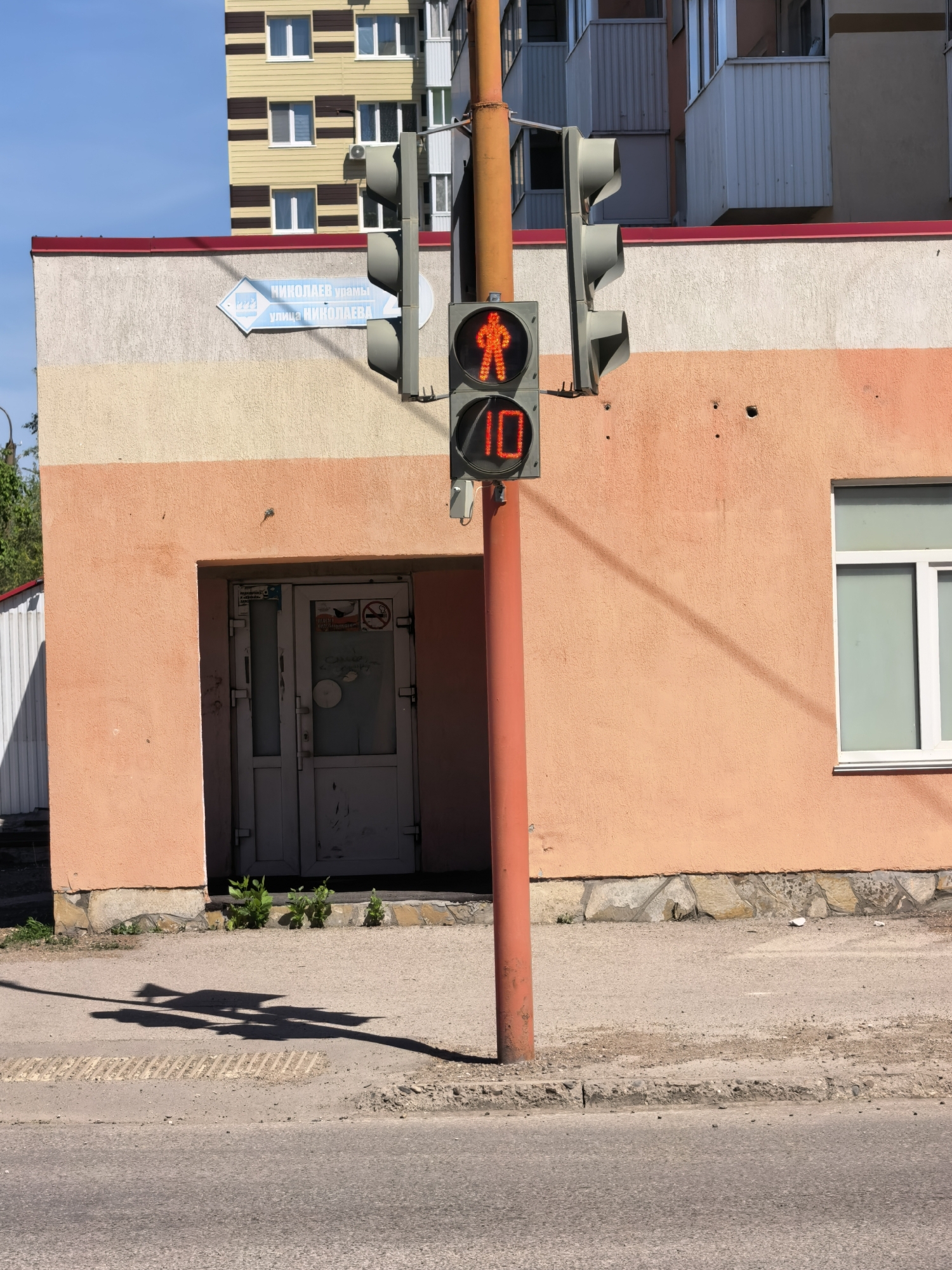 The image shows a pedestrian traffic signal in Sterlitamak, Russia. The signal displays a red pedestrian symbol and a countdown of 10 seconds. It is mounted on a red pole in front of a one-story building with a peach-colored facade. A street sign indicating "Nikolaev Street" is visible on the building's upper left side. The building has a gray door and a white-framed window. Part of a multi-story building is visible in the background. The street is paved with asphalt. The shadow of the traffic light is cast on the sidewalk.