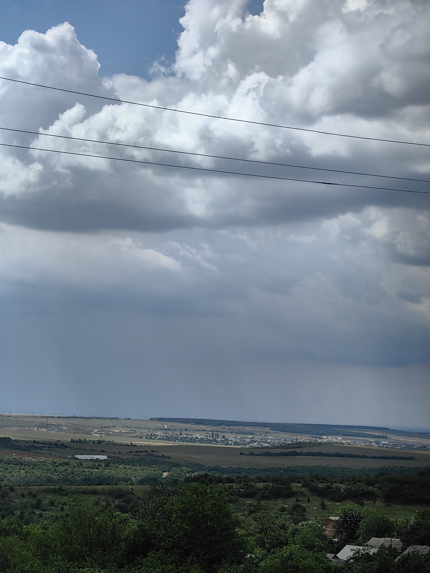 A wide shot captures a landscape under a dramatic sky with large, dark storm clouds partially obscuring a brighter sky above. In the foreground, dense green trees and a few rooftops of houses are visible, suggesting a rural or semi-rural setting. Beyond the trees, a flat expanse stretches out, dotted with various structures, including what appears to be a town or village in the distance. The terrain is varied, with rolling hills and open fields. Three power lines stretch horizontally across the upper portion of the image, creating a visual division between the sky and the landscape. The overall impression is one of an approaching storm over a populated area.