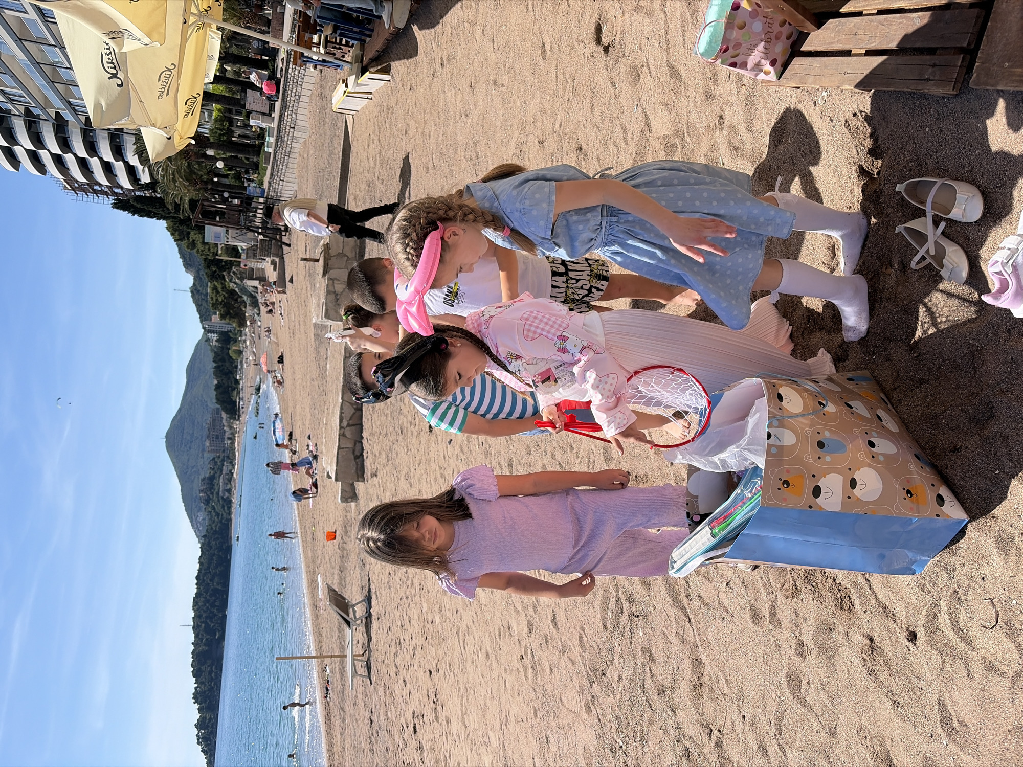 The image depicts a sunny day at Rafailovići beach, Montenegro. The view includes the Adriatic Sea, a distant mountain, and a beach crowded with people enjoying the day. In the foreground, a group of four children, three girls and one boy, are gathered around a gift bag placed on the sand. The girls are dressed in party attire: one wears a light blue polka-dot dress, another a pink kimono, and the third a purple outfit. The boy is wearing a striped shirt. The gift bag has a blue bottom and a brown top with images of bears. Near the group are a pair of white shoes and two shopping bags, one decorated with bears and the other in light pink. Umbrellas and buildings line the edge of the beach.