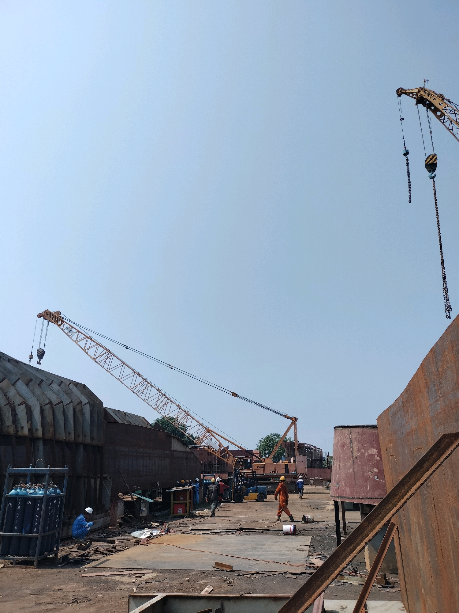 This is an outdoor image of a shipyard in Batam, Indonesia, under a clear, pale blue sky. In the foreground, to the left, a rack of multiple blue gas cylinders is visible. A person in a blue uniform is crouching next to them. In the midground, several workers are present, some wearing hard hats. One worker in an orange jumpsuit is walking across the frame. A yellow forklift is visible behind them. Two large industrial cranes dominate the scene, one in the midground stretching from left to right, and another larger one in the background on the right. The shipyard is filled with large, rusty metal structures, likely sections of ships under construction or repair. The ground is dirt and gravel, with scattered metal debris.