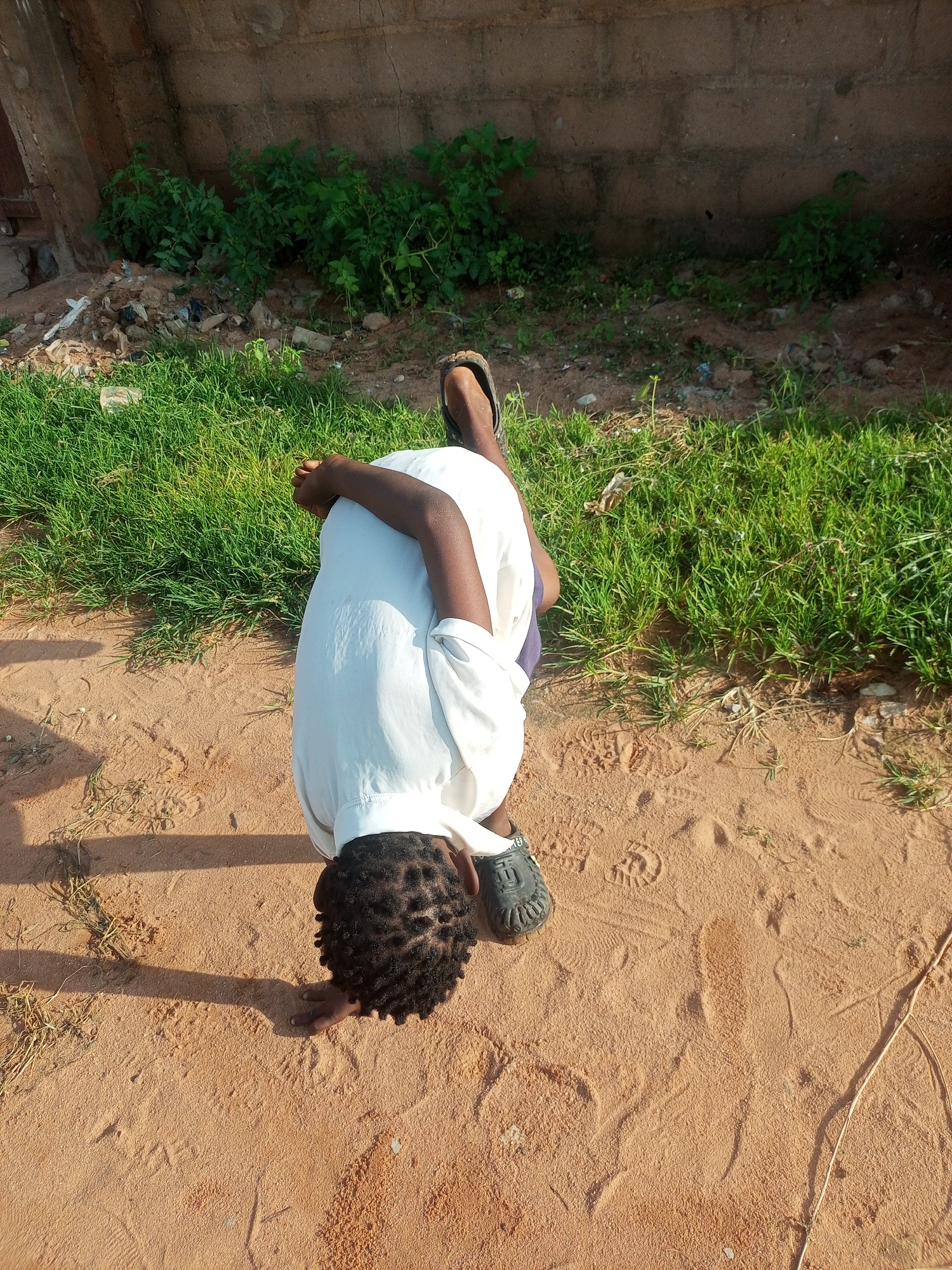 A person is lying prone on sandy ground in Tempure, Nigeria. The person is wearing a white t-shirt and dark shorts. Their head is bowed, and their hands are clasped together in front of them. Green grass is visible behind the person, and a rough concrete wall forms the background. Scattered debris is present in the grass and along the base of the wall. The ground is mostly bare sand with some footprints visible. The lighting suggests it is daytime, with shadows cast on the sand.