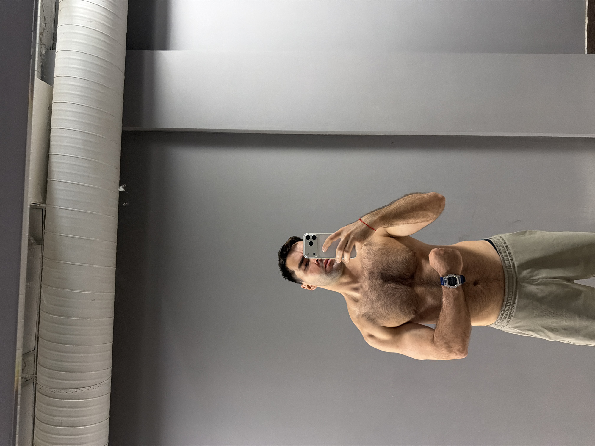 A shirtless man is taking a selfie in a mirror. He is wearing light-colored pants and a watch on his left wrist. The background is a gray wall and ceiling with a large, white ventilation pipe on the left side of the frame. There is no location in Krasnoyarsk, Russia, evident in the image.