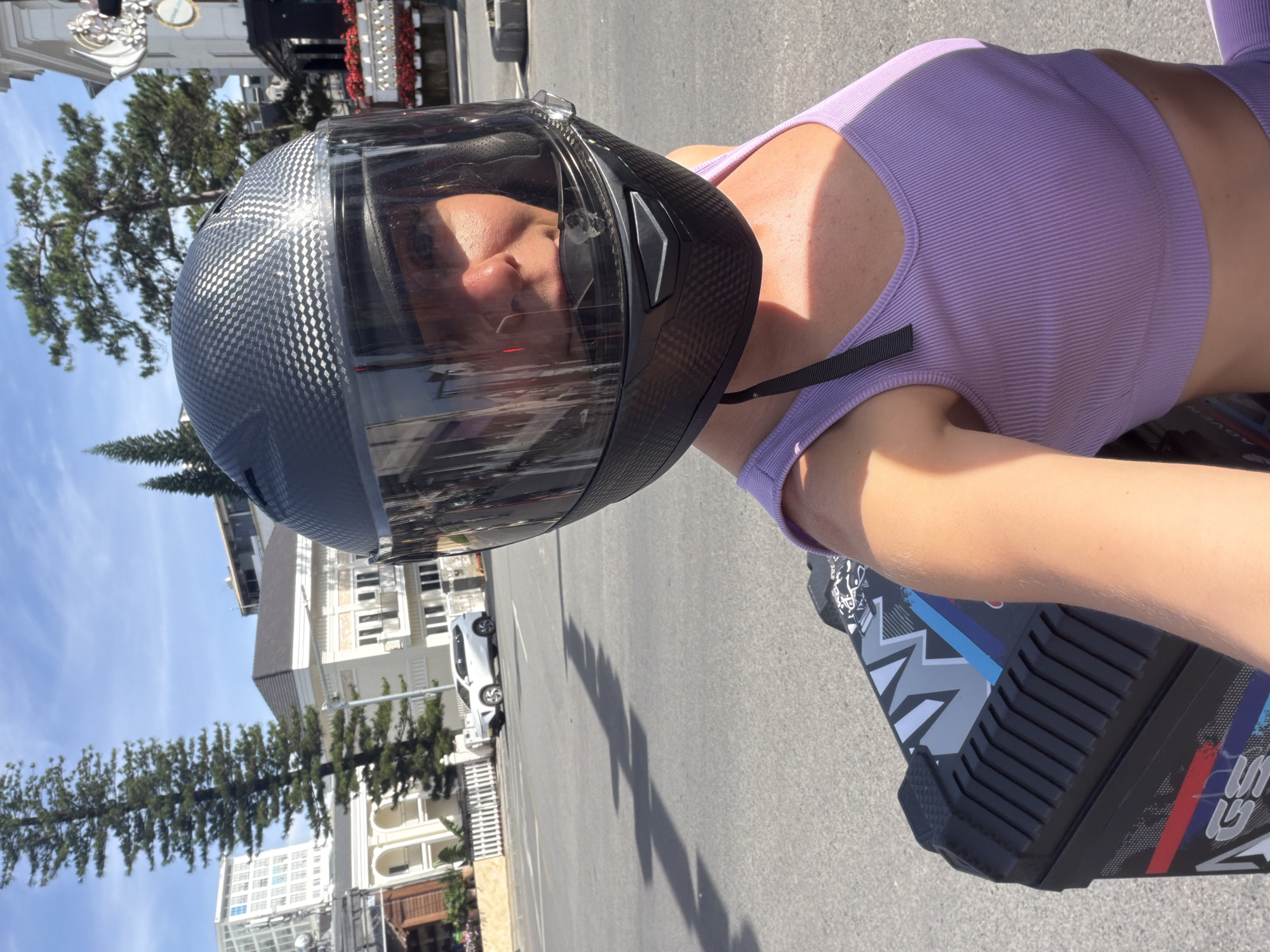 A woman wearing a black motorcycle helmet with a reflective visor and a light purple tank top is taking a selfie outdoors. She is positioned in the foreground, with a black storage box featuring a graphic design visible to her left. Behind her, a street scene unfolds with a white SUV parked on the side of the road and several buildings in the background, including white structures with arched windows and balconies, and a taller building with a modern facade. Pine trees and a clear blue sky with scattered clouds complete the environment. The scene appears to be in Da Lat, Vietnam, identifiable by the architectural style and foliage typical of the city.