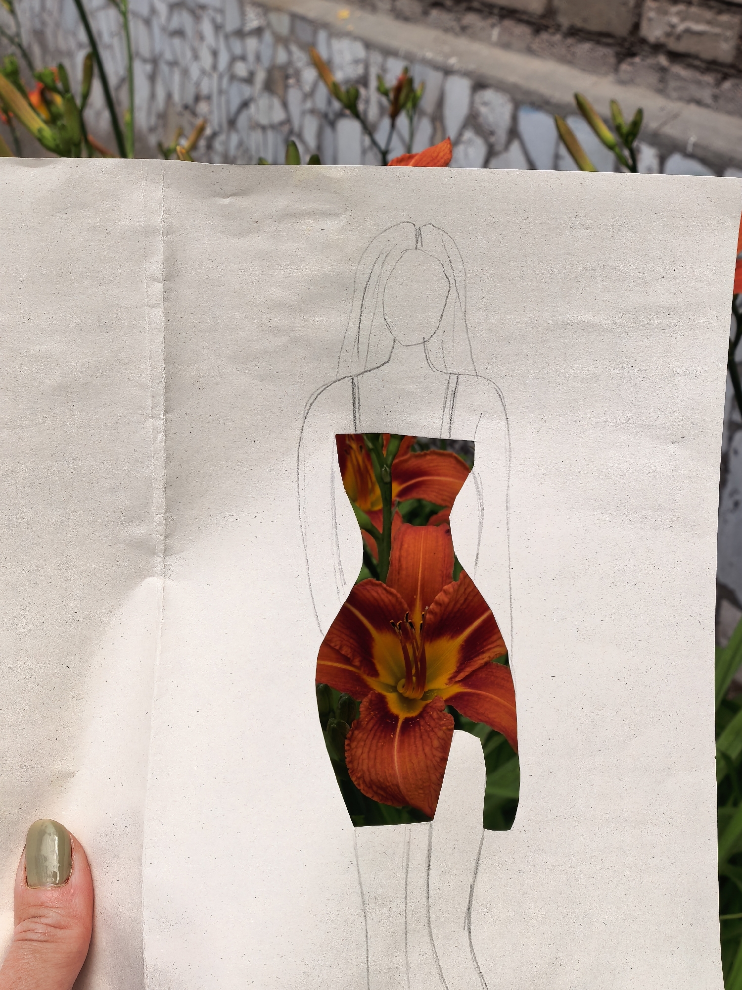 The image shows a close-up of a piece of paper with a rough sketch of a person's silhouette. The torso area of the silhouette has been cut out and replaced with a photograph of vibrant orange lilies. The paper is held by a person's thumb, which is visible in the lower left corner, with a light green nail polish. In the background, out of focus, there is a wall made of light-colored stones and some greenery, likely foliage and flower buds. The image does not provide any information about the location in the city of Sterlitamak, Russia.