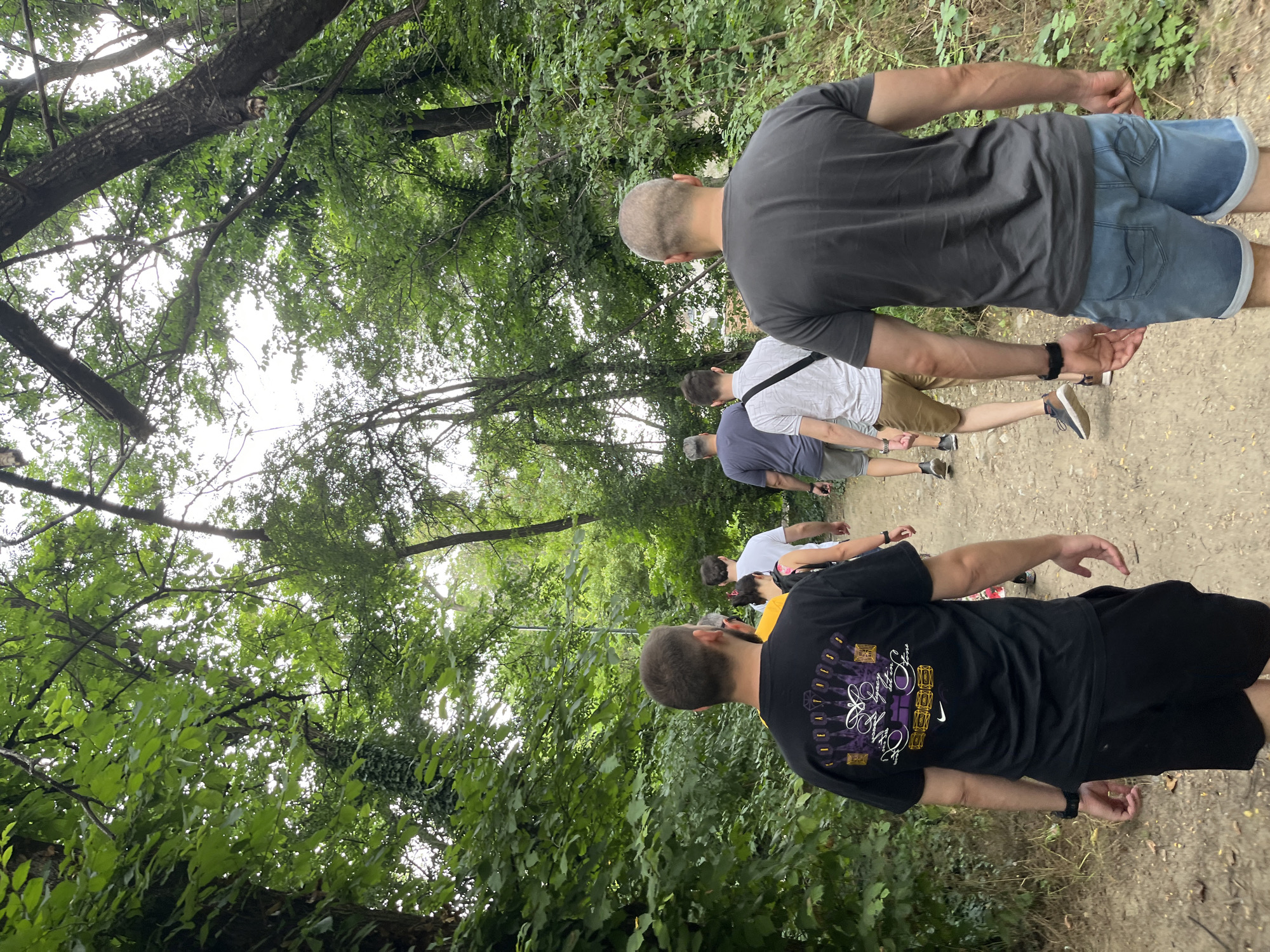 A group of six individuals, mostly male, walks on a dirt path through a wooded area. The path is flanked by lush green foliage and trees. The individuals are walking away from the camera, following the path. The sky is overcast and visible through the tree canopy. The scene appears to be an outdoor natural setting within the city of Plovdiv, Bulgaria, possibly a park or a forest trail.