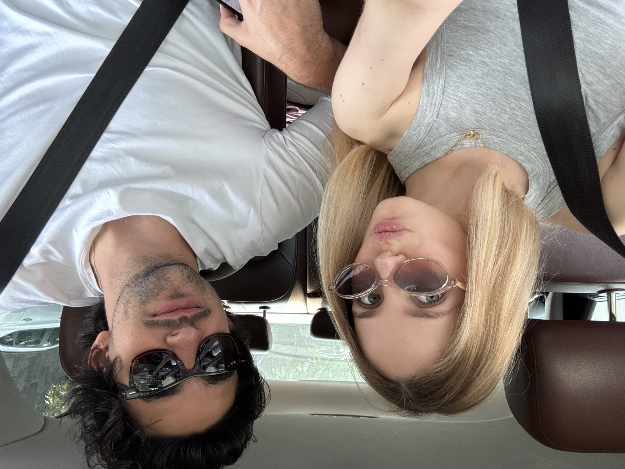 Two individuals, a male and a female, are positioned within the interior of a vehicle, both wearing seatbelts. The male is located on the left, wearing a white t-shirt and dark sunglasses. The female is on the right, wearing a grey tank top and round, rose-tinted sunglasses. The photo appears to be taken upside down, possibly by one of the occupants. The interior of the car has dark brown leather seats. Outside the car, a glimpse of trees and other vehicles suggests an urban environment. Without additional data, a specific location cannot be verified, but it is understood to be in Mexico City, Mexico.