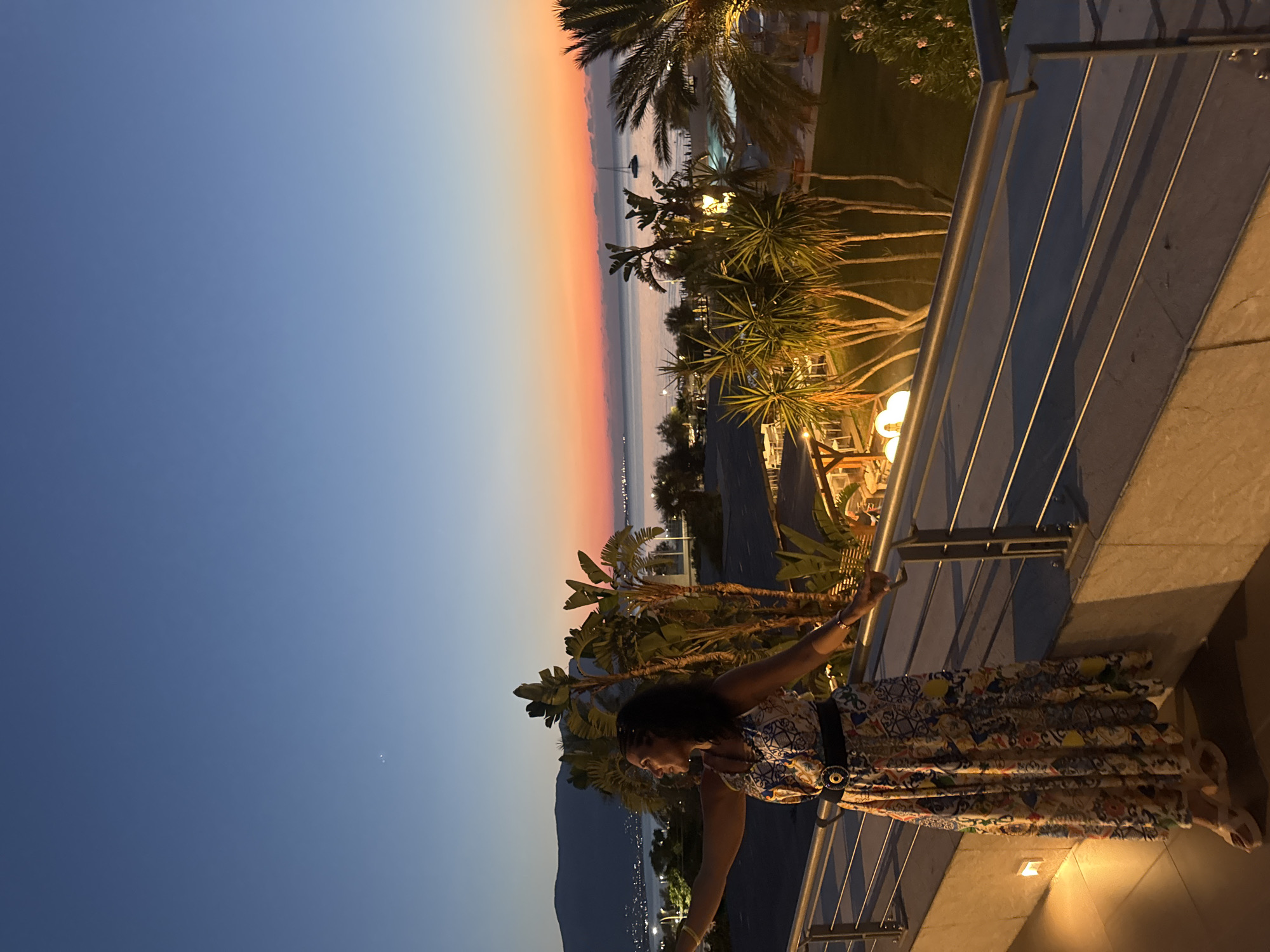 The image depicts a woman standing on a balcony overlooking a coastal town at dusk. The woman is wearing a long, colorful patterned dress and a black belt with a large, circular buckle. She is looking to her left with her arms extended. Palm trees and other lush vegetation are visible on the balcony and in the background. The sea is calm, with a sailboat anchored in the distance. The sky transitions from a deep blue at the top to vibrant shades of orange and pink near the horizon, indicating sunset or twilight. In the far distance, mountains are silhouetted against the sky. The balcony features a metal railing and ambient lighting, suggesting a hotel or residential building. The town lights are beginning to twinkle along the coastline. The overall impression is one of tranquility and beauty.