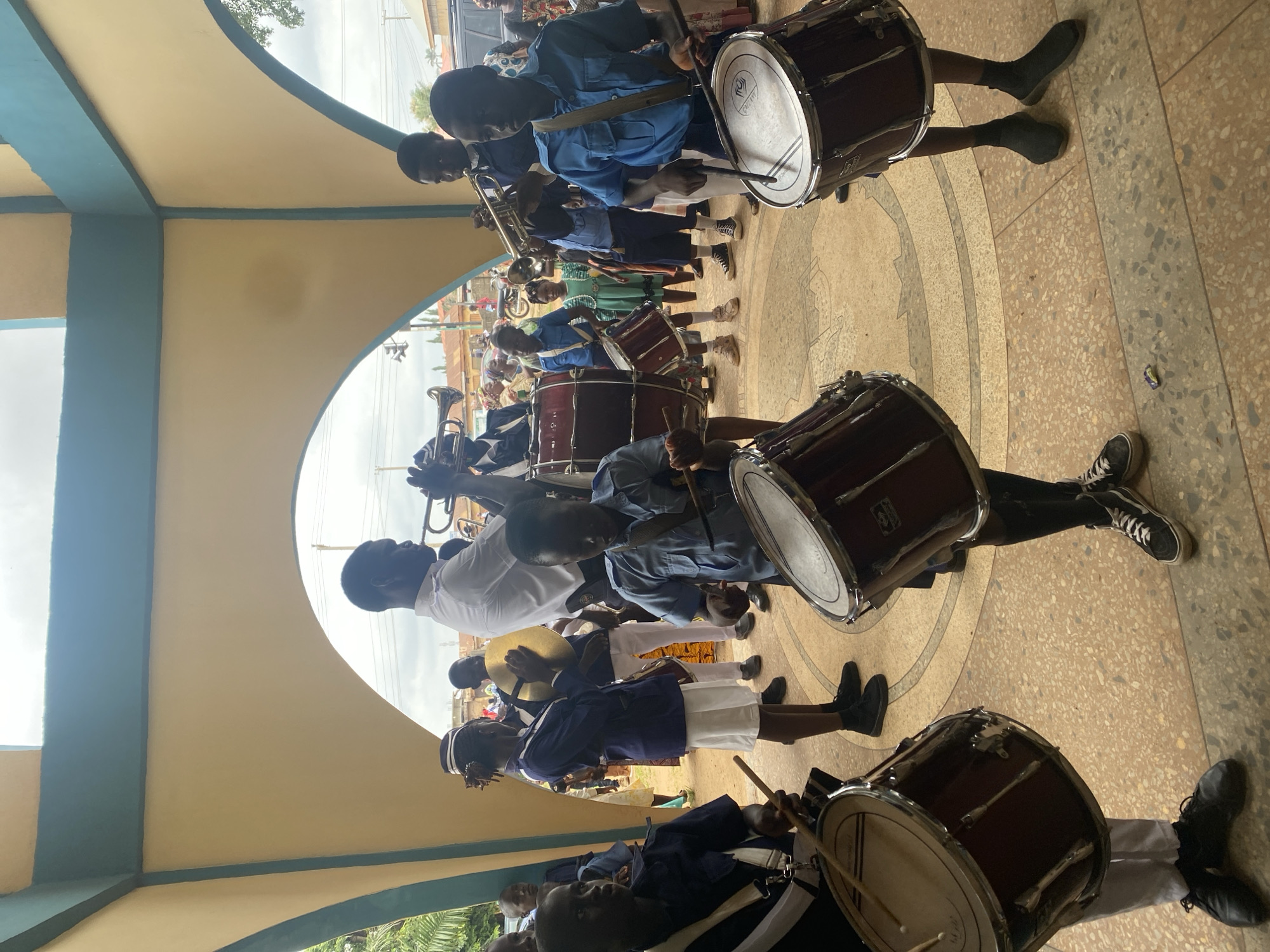 A band comprised of young people is performing inside of an arched walkway. The location is likely in Kafanchan B, Nigeria. The group consists primarily of drummers playing large drums. Other band members are playing horns. The band members are wearing uniforms. The walkway is painted in light blue and yellow. The ground is tiled.