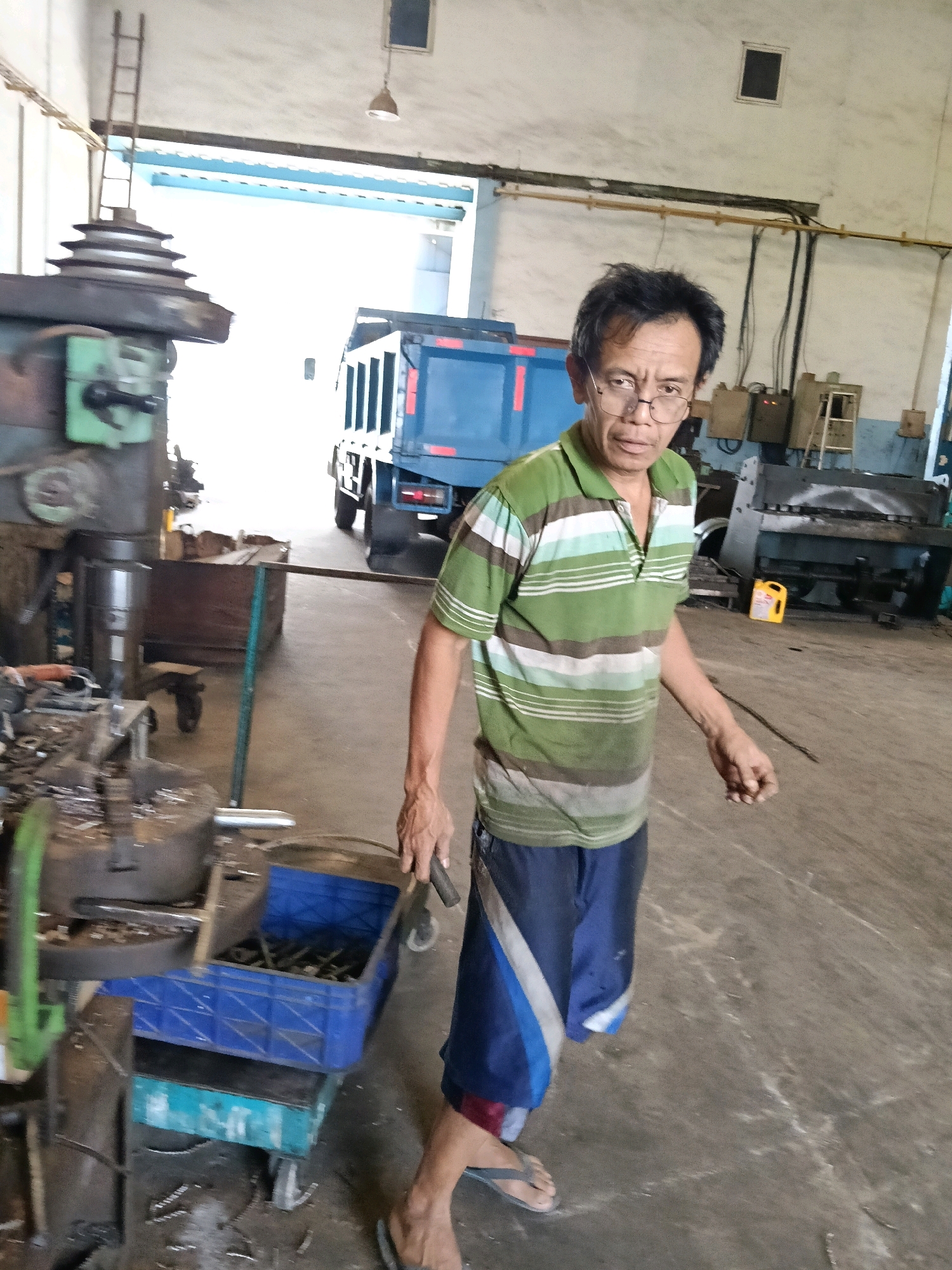 A middle-aged Asian man, wearing glasses, a striped polo shirt, and blue shorts, stands in a workshop. He is facing the camera with his right arm extended. In the background, a blue dump truck is parked, and various industrial machines are visible. The floor is concrete, and the overall environment suggests a busy work area. The man is likely a worker in this Indonesian city.