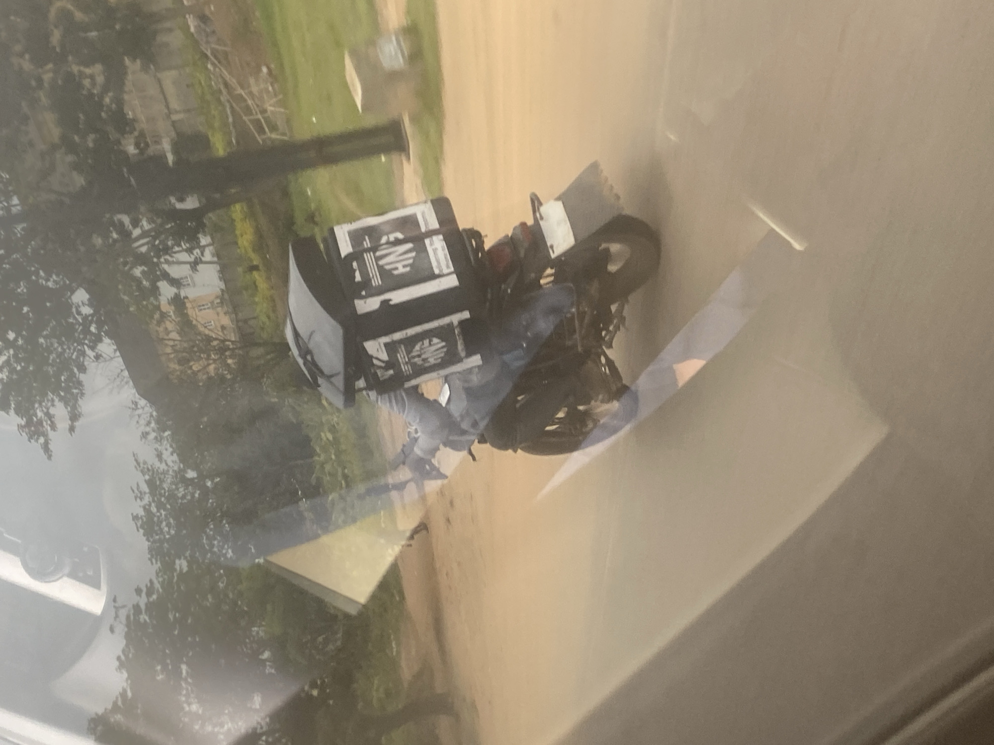 A delivery motorbike, possibly a courier or food delivery service, is parked on a dirt road, with a large branded delivery box attached to the back. The location appears to be in a somewhat undeveloped area, with trees and sparse vegetation. The image is taken through a window, causing reflections and obscuring the view. The precise location in Abuja, Nigeria is not discernible from the image.