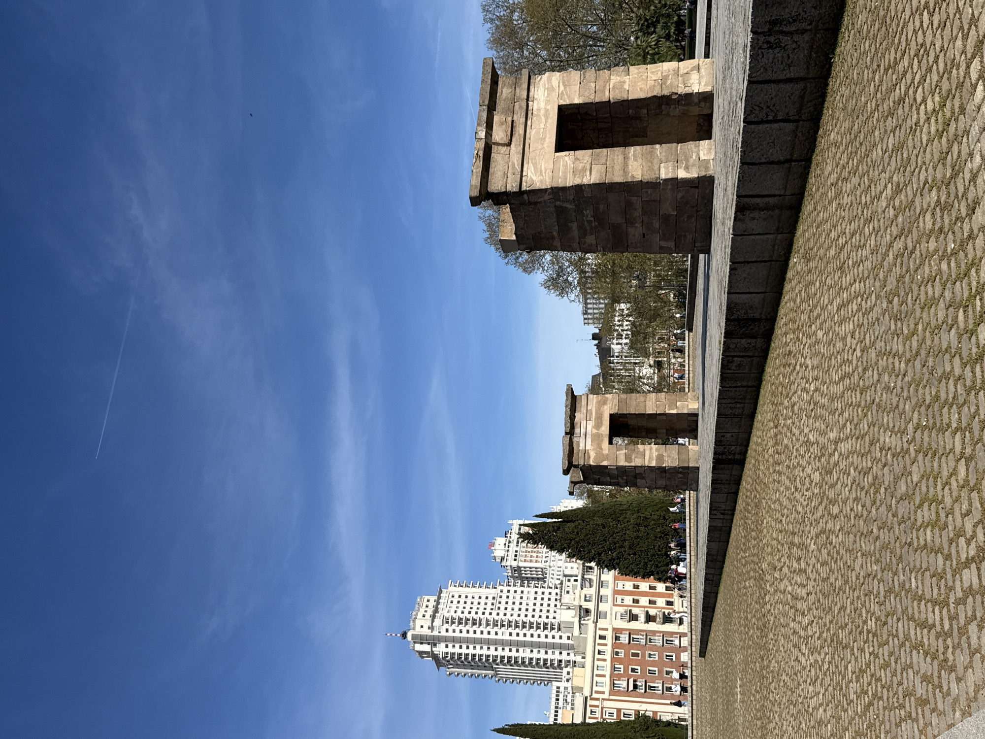 The image depicts the Temple of Debod in Madrid, Spain, under a clear blue sky with faint contrails. Two stone pylons of the temple are visible, with rectangular openings. Behind the temple, modern buildings rise, including a tall, white apartment building. A paved area stretches in the foreground. Trees are interspersed with the structures. The image is taken at an angle, giving a unique perspective of the temple and its surroundings.