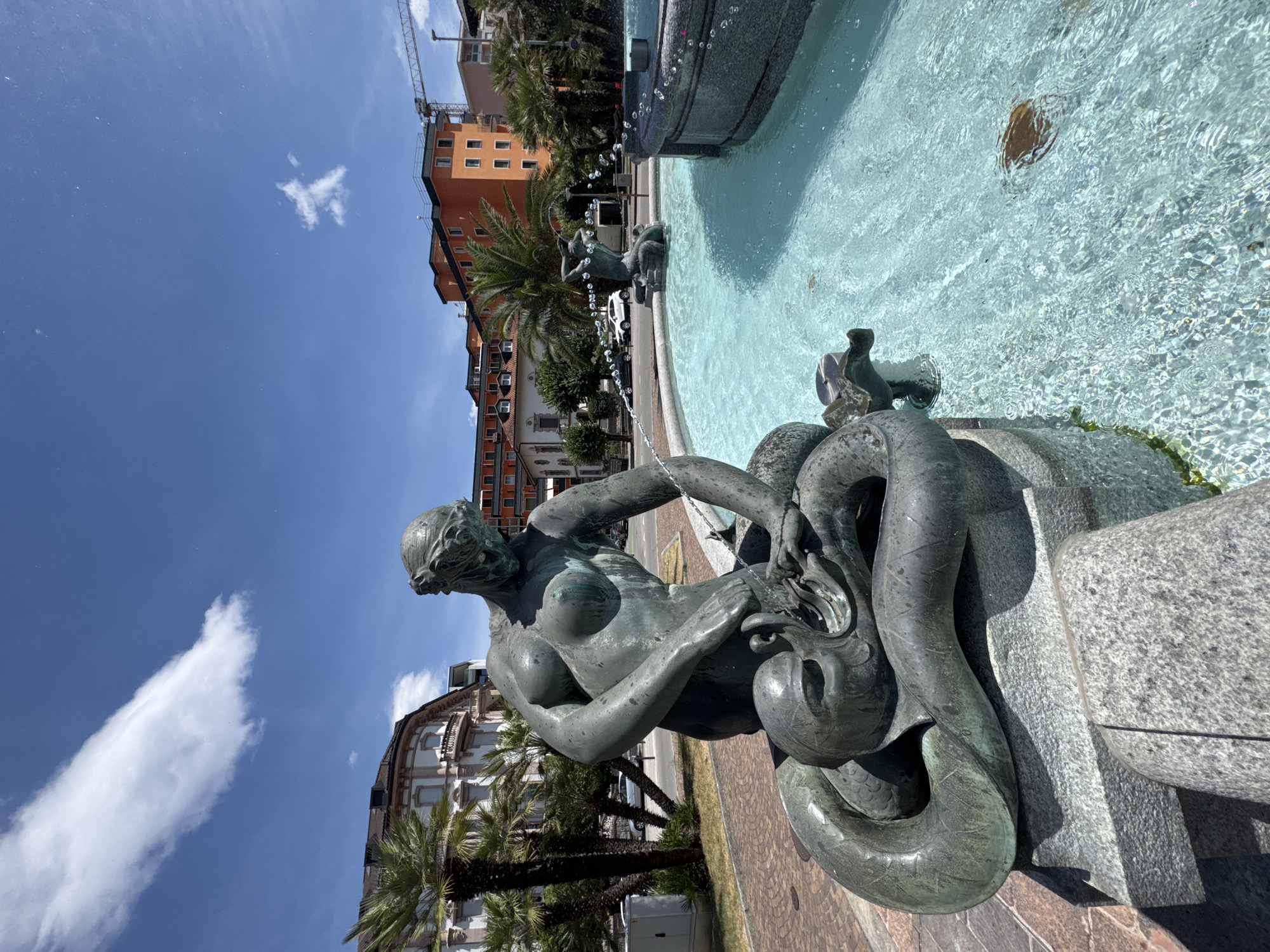 The image shows a bronze statue of a woman wrestling with a sea serpent, which is part of a fountain in Piazza Grande, Locarno, Switzerland. The statue is in the foreground, with the woman leaning over the serpent, and water spraying from her hand into its mouth. The fountain is filled with clear blue water, and there are palm trees and buildings in the background. The sky is blue with some white clouds. The statue is weathered and has a patina of green, indicating age. The overall scene is bright and sunny, suggesting a warm day.