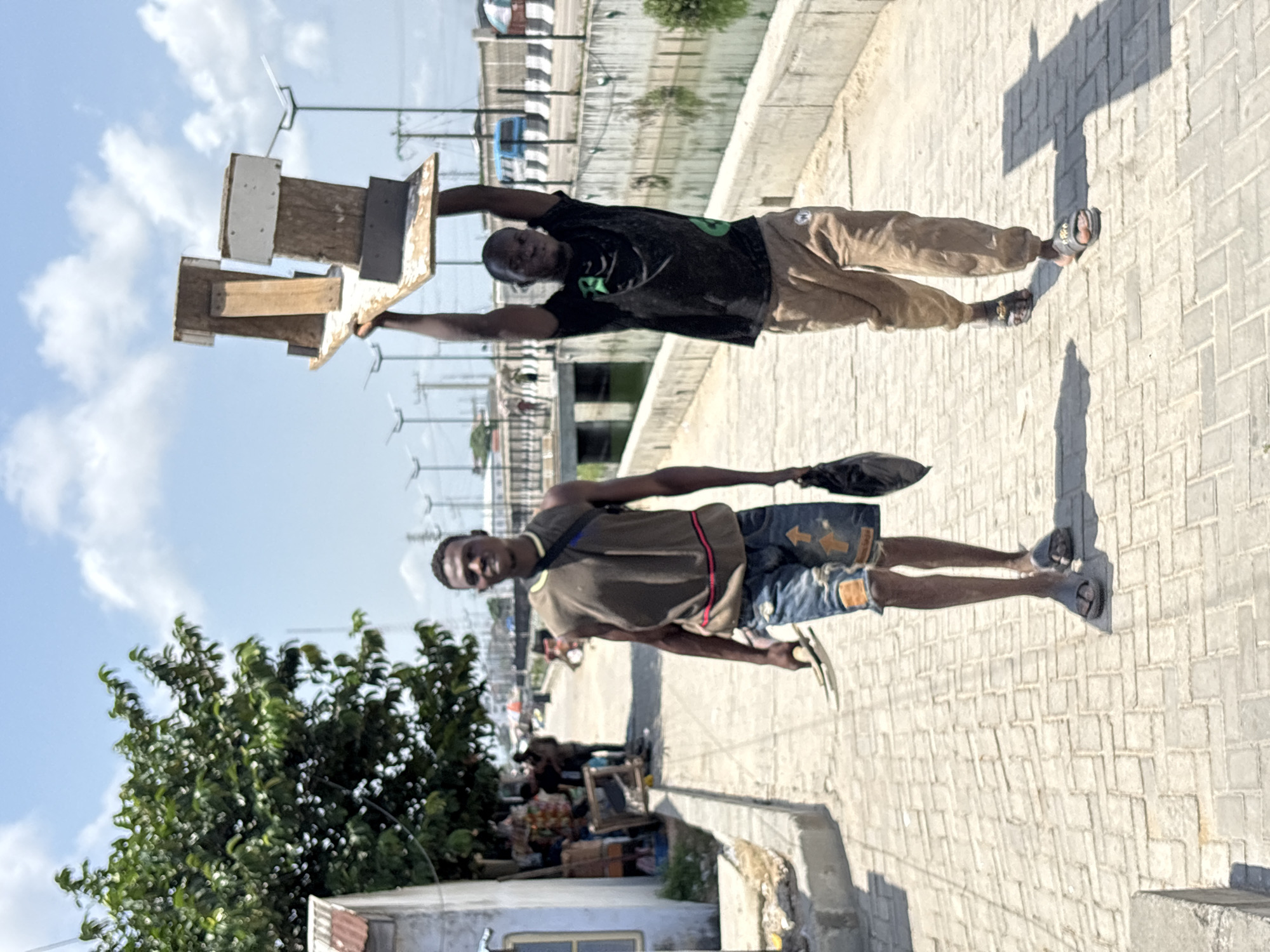 The photograph, taken in Igboefon, Nigeria, captures a vibrant street scene under a clear, bright sky with scattered white clouds. The image is originally oriented vertically but is presented rotated, showing two men walking on a paved sidewalk of interlocking bricks.
In the foreground, on the left, a man wears a brown t-shirt with horizontal red and black stripes, denim shorts adorned with yellow/orange arrow-like designs, and dark sandals. He carries a dark bag slung over his left shoulder and holds a flat, light-colored object in his right hand. He is looking towards the viewer with a slight smile.
Further back, on the right side of the sidewalk, a second man walks, balancing a tall stack of wooden structures on his head with both hands. These structures appear to be multi-level birdhouses or similar wooden boxes. He is dressed in a black t-shirt with a green design and light brown trousers, paired with dark sandals. His gaze is directed forward.
The bright sunlight casts long, distinct shadows of the men on the sidewalk, suggesting it is midday or early afternoon. To the immediate right of the sidewalk runs a concrete barrier, possibly alongside a canal or ditch. On the left side of the frame, a lush green tree partially obstructs the view. Beyond the tree and the men, the background reveals more of the urban environment, including what appear to be makeshift stalls or structures, a road, and in the distance, industrial poles or structures, possibly related to power lines or communication. The image depicts a daily life scene, likely involving street vending or the transport of goods.