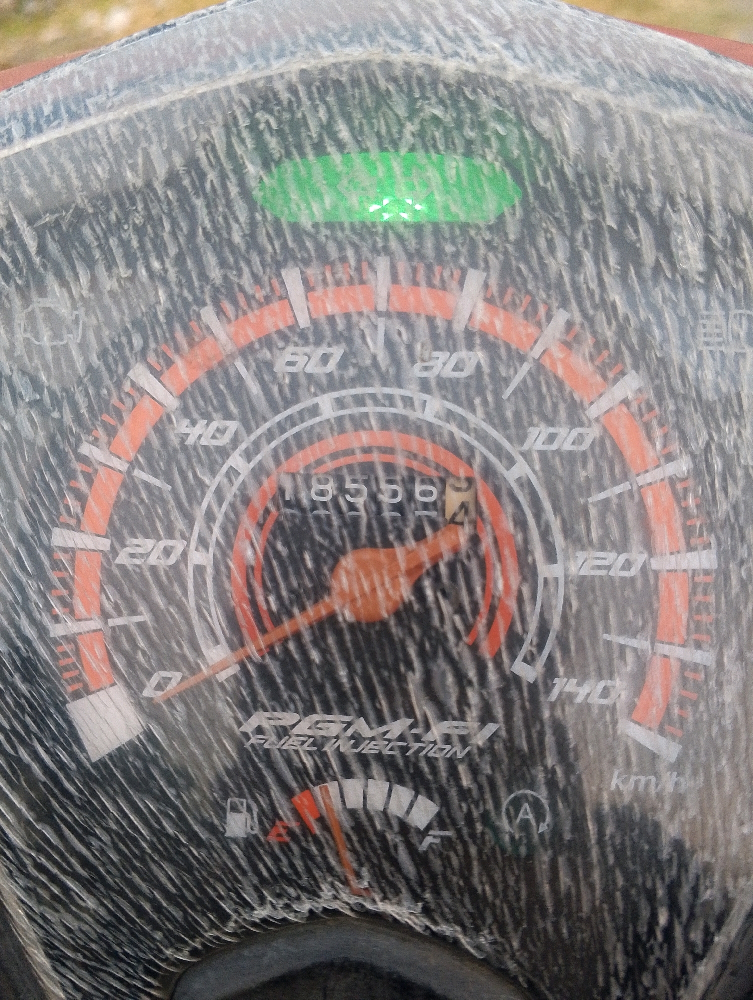 The image shows a close-up view of a dusty motorcycle speedometer. The speedometer has markings for speed in kilometers per hour, ranging from 0 to 140. The needle is currently pointing to approximately 56 km/h. There is also a digital display showing "56" and a fuel gauge indicator, which appears to be between half and full. A green indicator light is illuminated at the top of the speedometer. The environment appears to be outdoors, as indicated by the natural lighting and the texture of the surface beneath the motorcycle, which resembles soil or gravel. The image is captured in Pagiyanten, Indonesia.