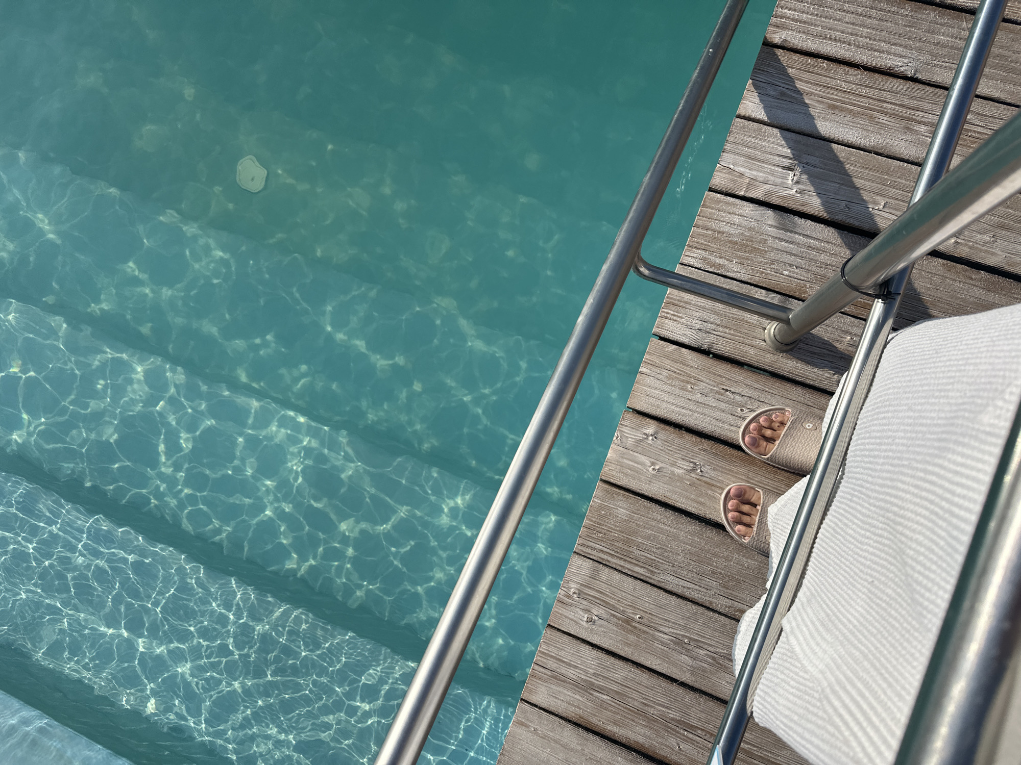 The image captures a top-down view of a person's feet and lower legs, wearing light-colored slides, resting on a wooden deck next to a swimming pool. The pool water is a clear turquoise color, with visible sunlight refracting on the submerged steps and a light-colored object, possibly a pool cleaner, at the bottom. A metal railing runs along the edge of the deck and the pool. The wooden deck planks are weathered and laid diagonally. The scene appears to be a recreational area, potentially a private residence or a resort. There is no specific indication of the location being in Yaroslavskaya, Russia.