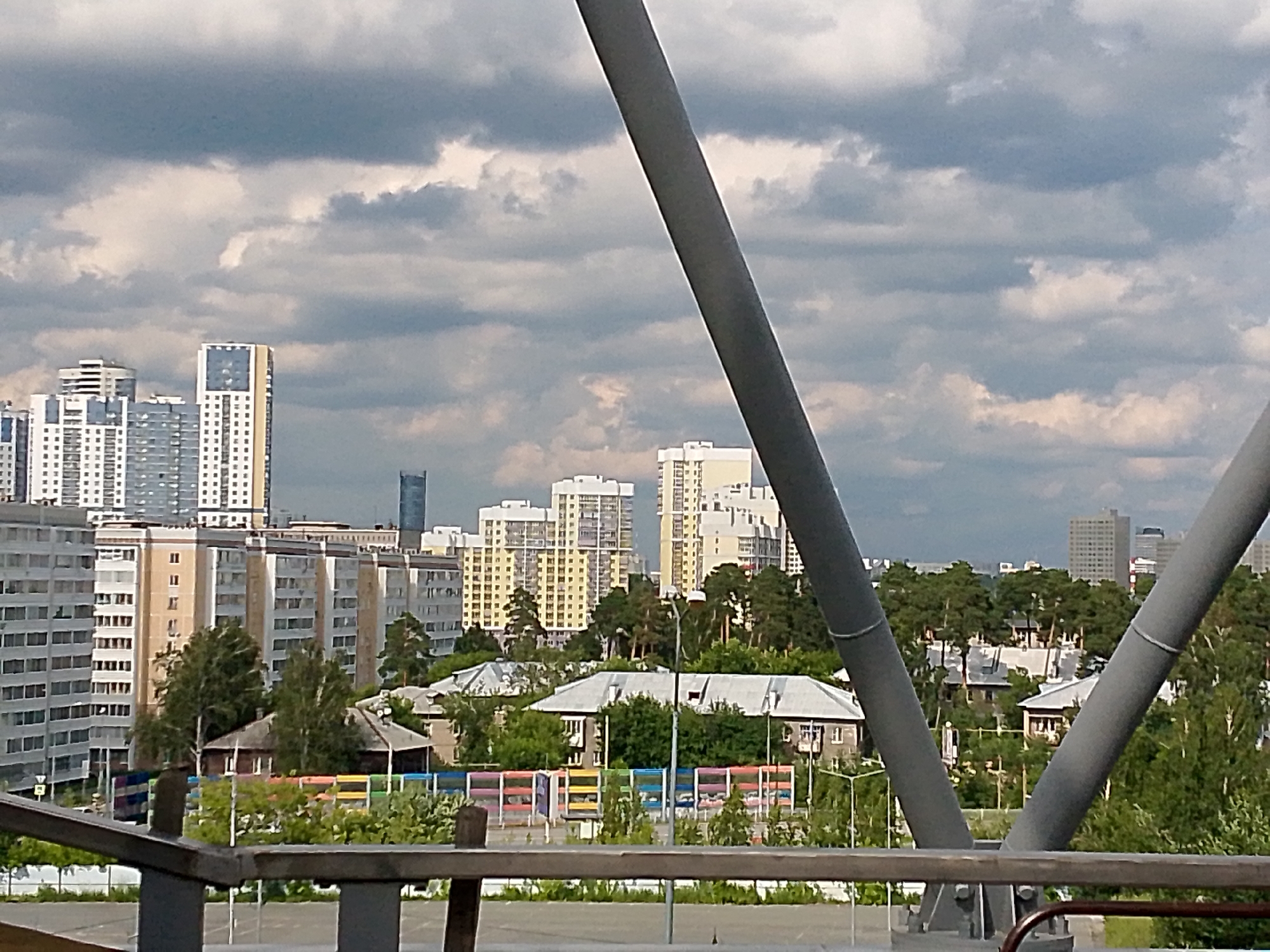 An exterior medium shot captures a city skyline dominated by high-rise buildings and a cloudy sky. The foreground is partially obstructed by large, gray metal beams, likely part of a bridge or overpass. Below the beams, a railing runs horizontally across the bottom of the frame.
In the mid-ground, a dense cluster of trees partially obscures a collection of lower-rise buildings with pitched roofs. These houses appear to be of older construction, with some featuring visible windows and chimneys. A colorful, abstract fence or barrier with horizontal bars in red, yellow, blue, and green runs in front of these houses.
The background showcases a mix of modern architecture. Several tall apartment buildings, some with yellow facades and white window frames, and others with gray and white exteriors, punctuate the skyline. A slender, dark skyscraper is visible to the left of the frame. The sky is filled with dramatic, dark gray clouds, suggesting an overcast or potentially stormy day. The overall scene depicts a transition from a more established residential area to a growing urban landscape.