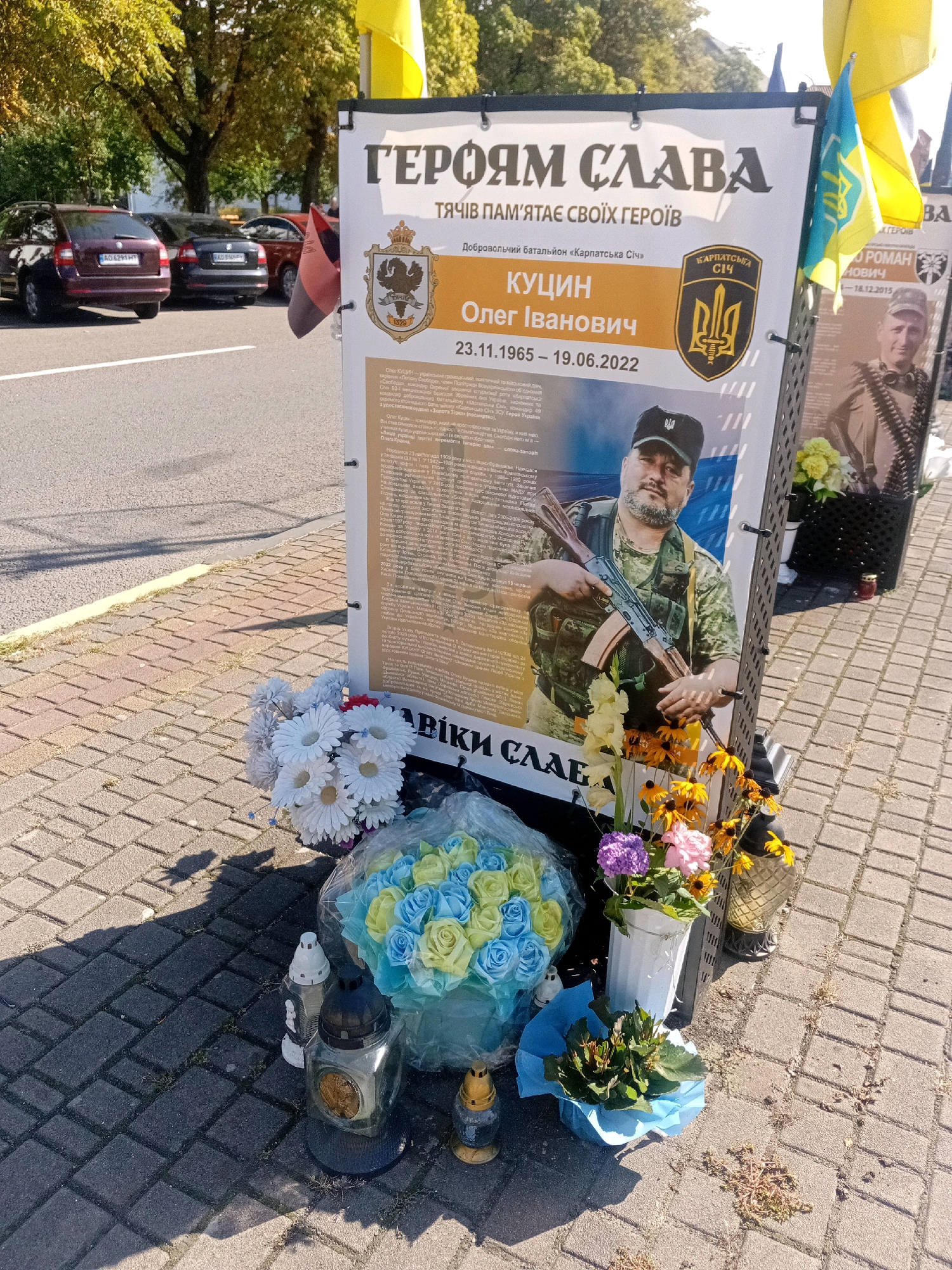 A memorial display for Oleh Ivanovych Kuznetsov, a Ukrainian soldier, stands on a paved sidewalk in Tiachiv, Ukraine. The display features a large poster with Kuznetsov's portrait, his dates of birth and death (November 23, 1965 – June 19, 2022), and biographical information. Two Ukrainian flags and a flag with a trident emblem are attached to the display. Floral arrangements and lit candles surround the base of the memorial, with a bouquet of blue and yellow roses placed prominently in front. In the background, cars are parked along a road, and trees with green and yellow foliage suggest an outdoor setting. The overall scene is solemn, indicating a tribute to a fallen hero.
