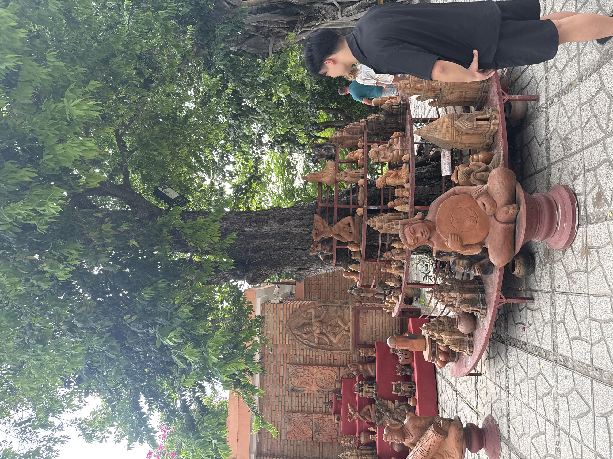 A young Asian man in a black t-shirt and shorts stands facing right, his left hand in his pocket, observing a display of terracotta sculptures outdoors. The sculptures are arranged on tiered shelves under a large tree, with a brick wall and building in the background. A few other people are visible in the distance, walking away from the viewer. The ground is paved with light-colored tiles. This scene appears to be at the Pottery Village in Nha Trang, Vietnam, a place known for its traditional pottery and art.