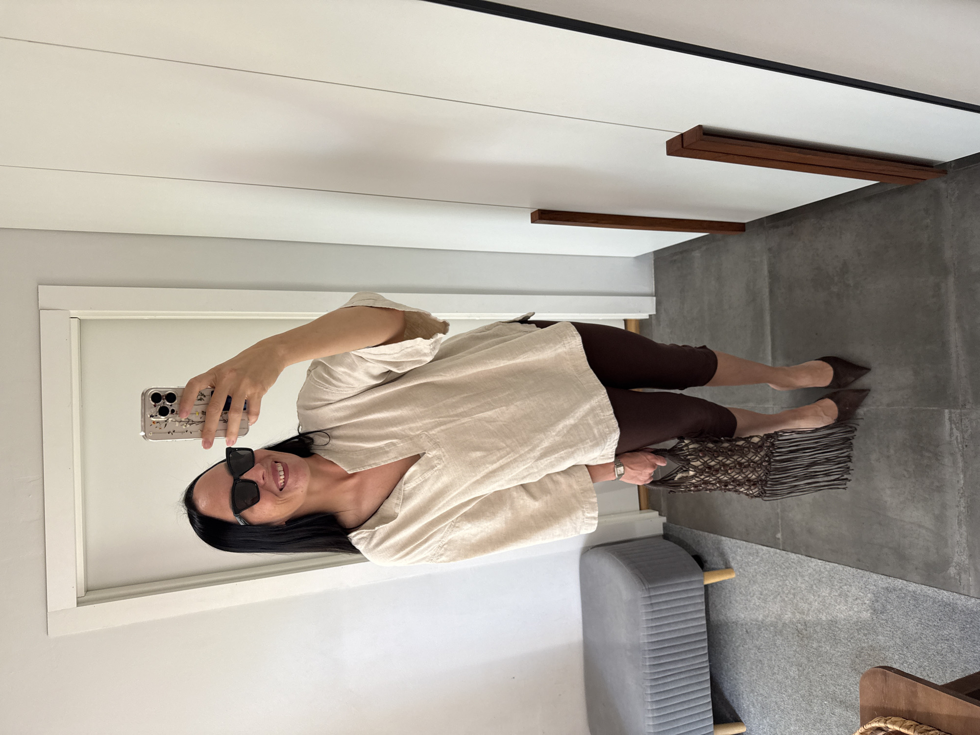 A woman stands in a hallway, taking a selfie in a mirror. She is wearing sunglasses, a light-colored linen tunic, dark brown capri pants, and pointed-toe heels. She is carrying a small macrame bag with fringe. To her left, a gray upholstered bench with wooden legs sits on a gray rug. The floor is tiled in a dark gray. The walls are white, and a white door is visible on the left. The scene appears to be indoors, possibly in a home or a retail space. There is no indication of the city of Chaikovsky, Russia.