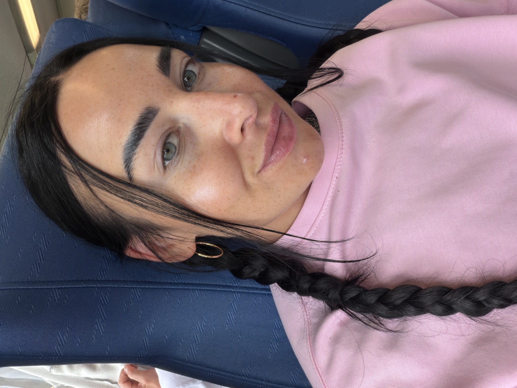 The image shows a close-up, selfie-style shot of a young woman with dark hair in braids, wearing a pink sweatshirt. Her eyes are green and she has freckles on her face. She is seated on a blue upholstered seat, possibly on a train or bus, indicated by the texture of the seat material. Her left ear is visible, adorned with a gold hoop earring. The background is dominated by the blue seat and a sliver of a white garment on the left. There is no indication of the location being the city of Ушаки, Russia.