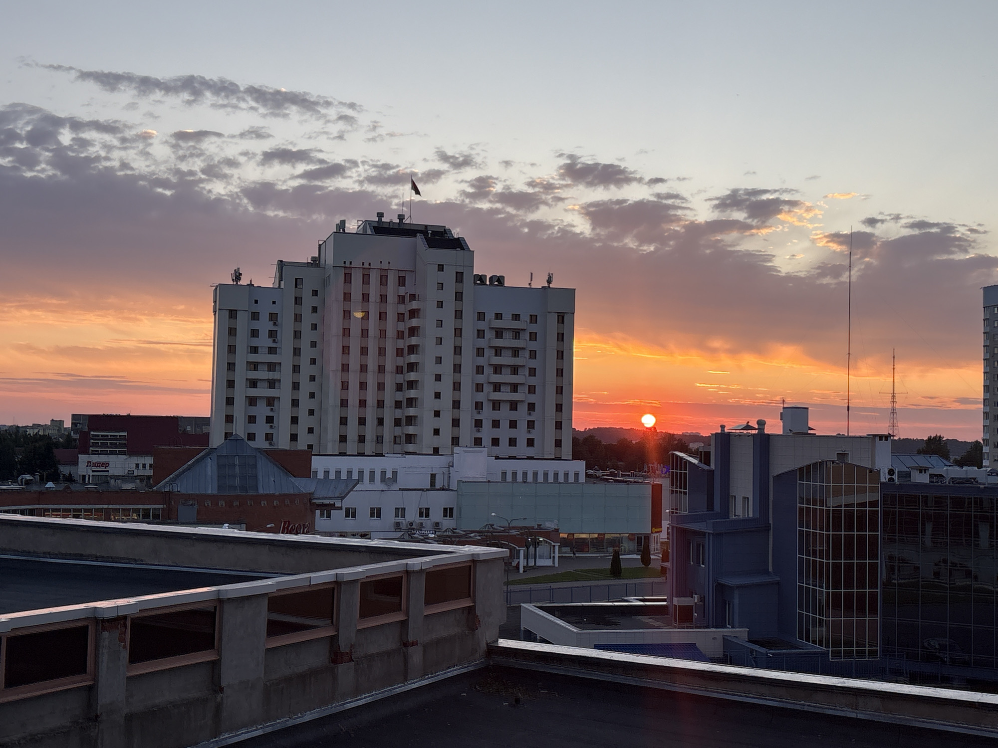 The image depicts an urban cityscape at sunset, with a bright orange sun low on the horizon, casting warm light and long shadows. In the foreground, a rooftop with a concrete barrier is visible. Dominating the mid-ground is a tall, multi-story white building with numerous windows, topped with a flag. Adjacent to it, a series of modern buildings with glass facades and varied architectural styles are present. In the background, more city structures and greenery can be observed, merging with the colorful sky. The scene is likely captured from an elevated vantage point within the city of Vitsebsk, Belarus, given the presence of a distinct Soviet-era style apartment block and modern office buildings. There are no people or animals visible in the frame. The overall atmosphere is serene and captures the transition from day to night.