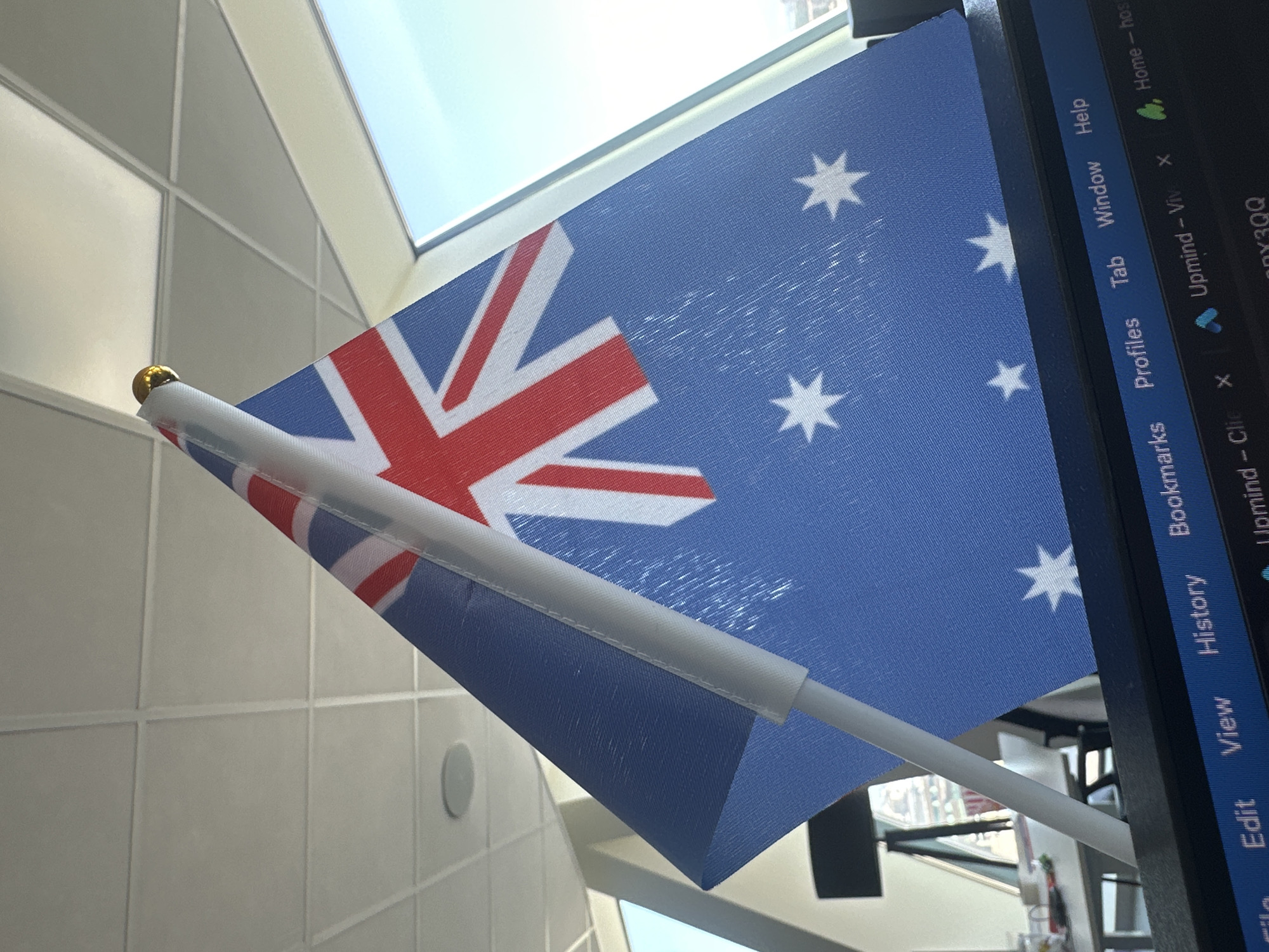 The image shows a small Australian flag placed on top of a computer monitor, against the backdrop of an office environment. The ceiling tiles and a window with a view of the sky are visible in the background. The computer monitor displays a web browser with tabs open. The scene appears to be an indoor office space. There are no people visible in the image. The specific location in the city of Sofia, Bulgaria, is not identifiable from the image alone.