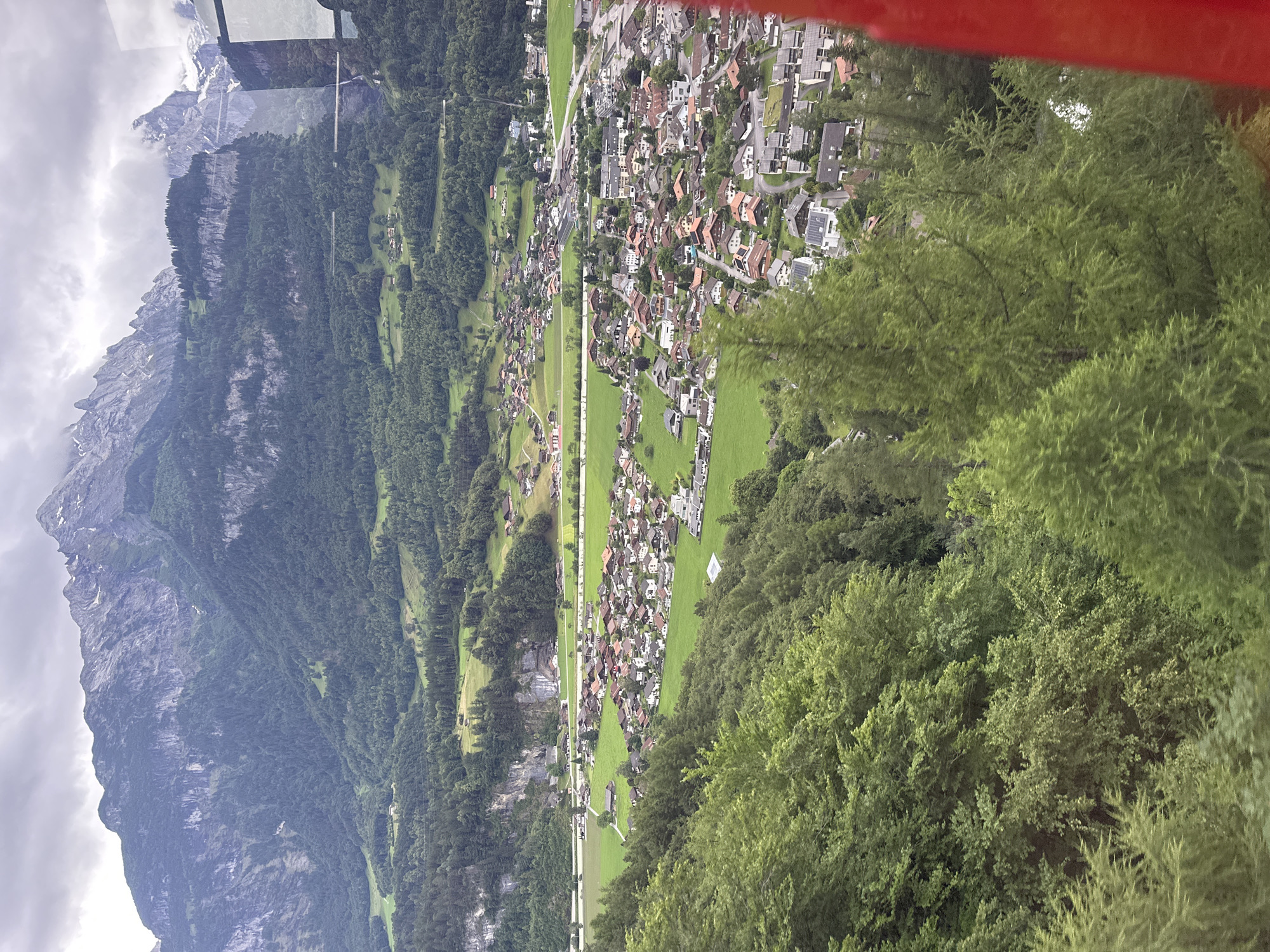 The image depicts a panoramic view of a town nestled in a valley, with imposing mountains in the background. The town is characterized by its red-tiled roofs and white-walled houses, densely packed together in some areas and more spread out in others. Green fields and forests surround the town, creating a stark contrast with the rugged, rocky peaks of the mountains. The sky is overcast, with dark clouds hinting at impending rain or a storm. A red structure, likely part of a vehicle or gondola, is visible in the foreground, suggesting that the photograph was taken from an elevated perspective. The overall impression is one of a serene, yet dramatic, natural landscape, with human settlement integrated into the environment. The specific location in the city of Hüsen, Switzerland, is identifiable by the characteristic architecture and the surrounding alpine scenery.