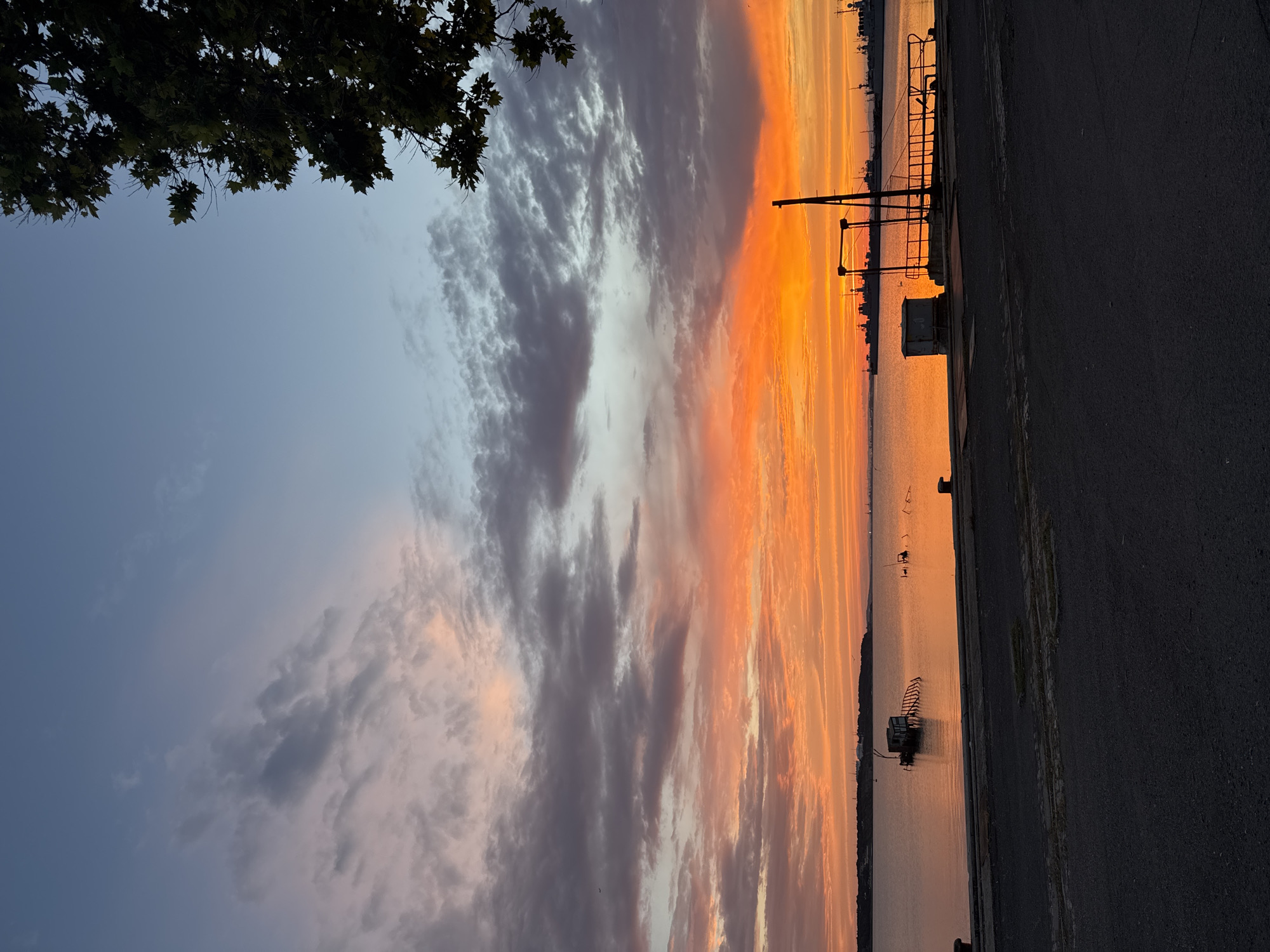 The image depicts a serene sunset over the sea, likely in Burgas, Bulgaria. The sky is ablaze with vibrant orange and pink hues, transitioning into softer blues and grays higher up. Wispy clouds are scattered across the horizon, catching the warm light of the setting sun. In the foreground, a paved area with what appears to be old railway tracks leads to a waterfront.
The water is calm, reflecting the colors of the sky, and shows some debris or structures partially submerged. A small, weathered hut-like structure with a leaning ramp sits in the water on the left side of the image. Further out, a series of darker shapes on the horizon suggest ships or a distant shoreline with industrial elements. A metal structure with railings, possibly part of a pier or dock, is visible on the right. A lone tree branch with green leaves frames the top right corner of the image. There are no people visible in the scene, and the overall atmosphere is tranquil and still.