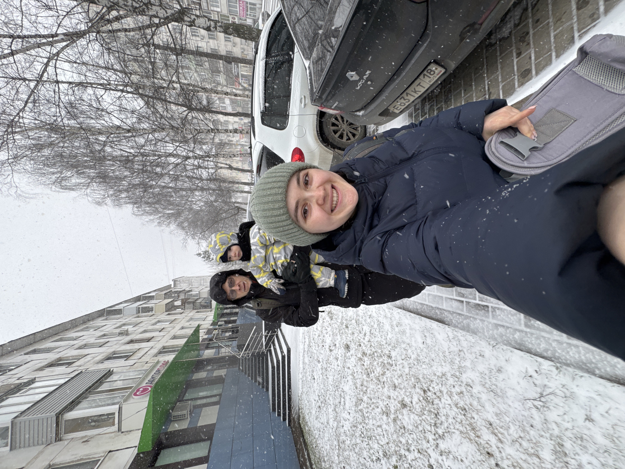 Three people are in the frame, standing in the snow. The person closest to the camera is a young adult wearing a beanie and a dark blue winter coat. Behind her are two others: one appears to be holding a child dressed in a yellow and white patterned coat with the hood up. The background features a cityscape with buildings, bare trees, and parked cars covered in snow. Based on the license plate "E631KT 18 RUS", the location is Izhevsk, Russia. It's snowing.