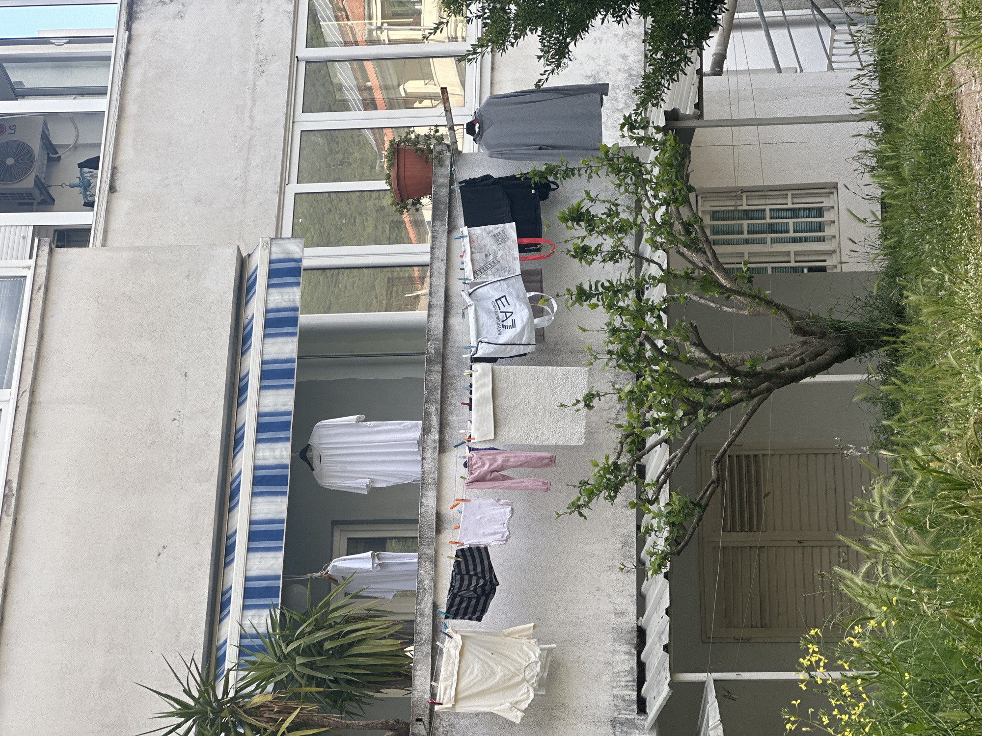 A building facade in Budva, Montenegro, features an exterior wall where clothes are hanging to dry. The wall is off-white and shows signs of weathering. A striped blue and white awning is visible, and several windows reflect the outside environment. A potted plant sits near one of the windows. Clothes of various colors, including grey, black, white, pink, and striped patterns, hang from a clothesline secured to the wall. A tree with green leaves grows in the vicinity, partially obscuring the view. A small patch of grass and yellow wildflowers are also present. An air conditioning unit is visible on one section of the building's facade. Shopping bags also hang on the line. The style of the architecture is typical of residential buildings in the Mediterranean region.