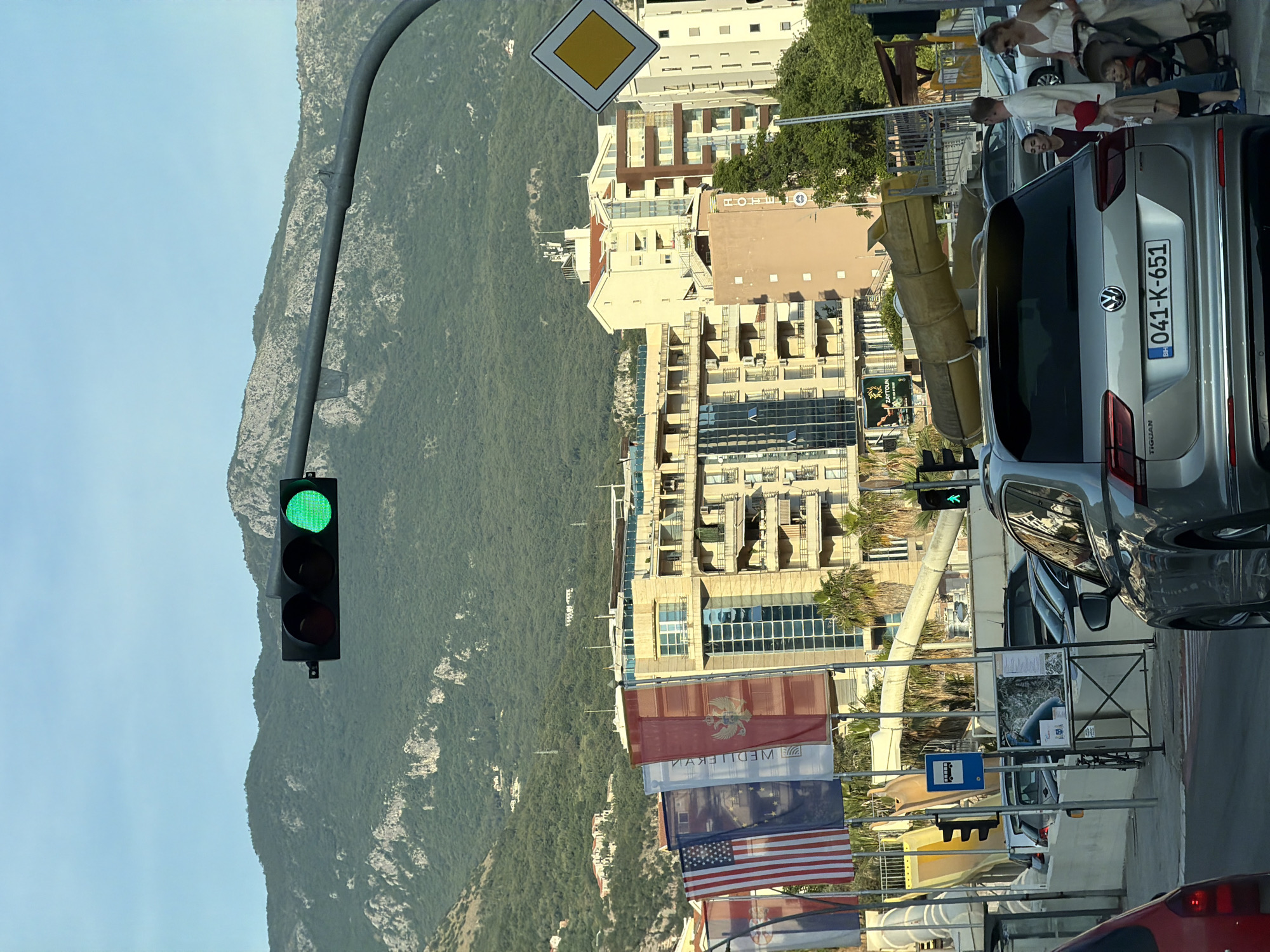 The image shows a street scene in Boreti, Montenegro. A silver Volkswagen Tiguan with Bosnian license plate 041-K-651 is parked on the side of the road. A woman pushing a baby stroller and a man standing beside her are visible near the car. Traffic lights are mounted on poles, both showing green. A mountain range is in the background, and buildings are on the right side of the street. Several national flags, including the United States flag, are hanging on poles along the sidewalk. A yellow diamond-shaped road sign is also visible.