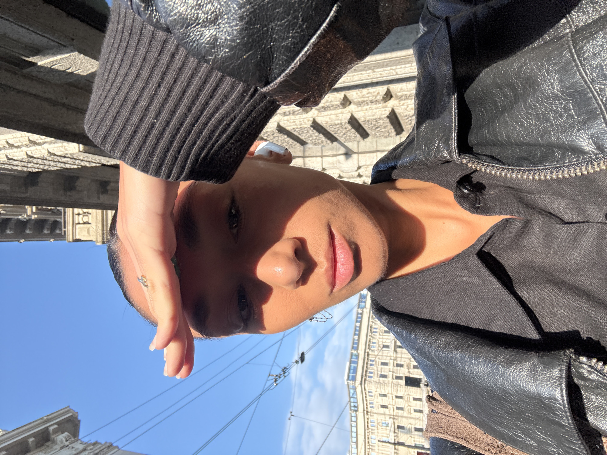 A selfie-style photograph features a person with brown skin, shielding their eyes from the sun with their left hand. The person is wearing a black leather jacket with a ribbed cuff on the sleeve. They have an earpiece in their left ear and a small ring on their left hand. The background reveals a cityscape, identifiable by the architectural style as Milan, Italy. Buildings with repeating window patterns and detailed stonework are visible, suggesting a European urban setting. Above the buildings, a clear blue sky with light clouds is visible. Power lines extend across the frame. The photo has been taken during daylight hours, as indicated by the brightness and shadows.