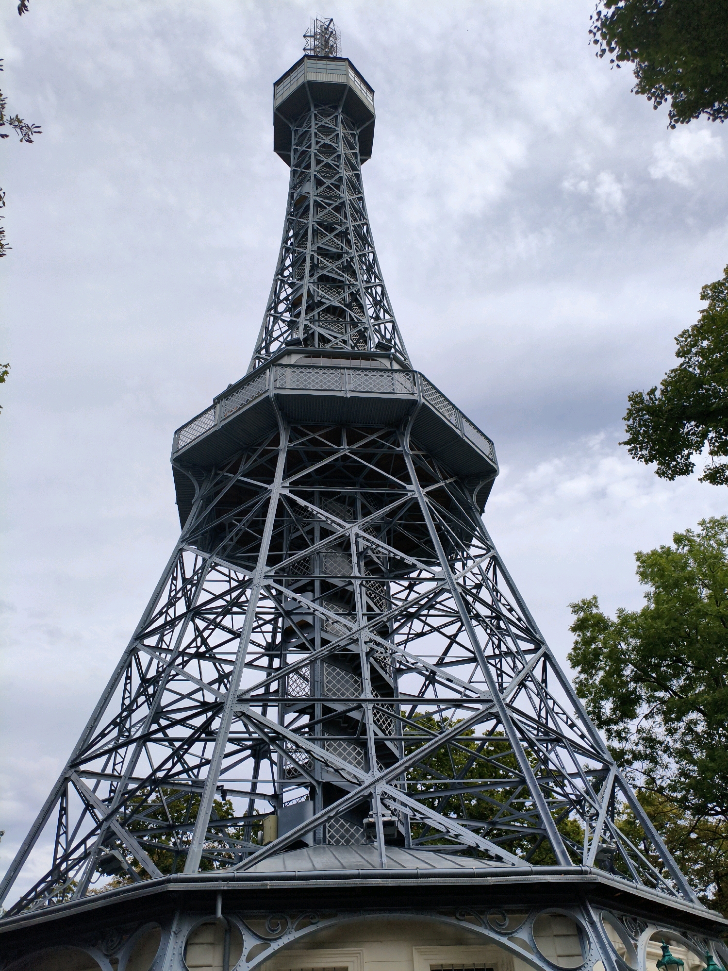 A low-angle, full shot captures the Petřín Lookout Tower, a steel lattice tower, against a cloudy sky. The tower, resembling a miniature Eiffel Tower, has a wide, conical base that narrows as it ascends to a rectangular observation deck and a smaller, segmented tower at the very top. The latticework construction of the tower is intricate, with diagonal and cross bracing visible throughout. A spiral staircase is partially visible within the tower's structure. Green trees are visible on the right and left edges of the frame, framing the tower. The base of the tower appears to sit on a white structure with decorative ironwork. No people are visible in the image. The scene is set in Prague, Czechia.
