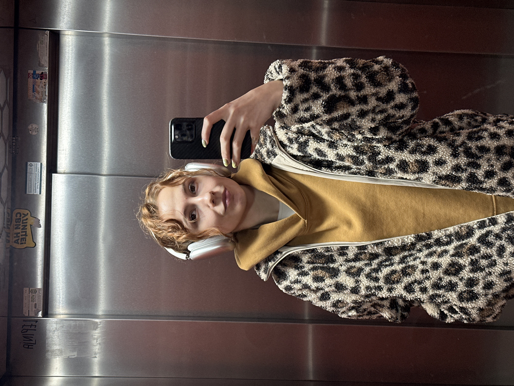 The image, rotated 90 degrees counter-clockwise, depicts a young person taking a selfie inside an elevator. The individual, positioned slightly right of the frame's center, has light brown or reddish wavy hair and wears white over-ear headphones. They are looking directly at the camera with a neutral expression.
The person is dressed in a mustard yellow hooded sweatshirt, layered under a textured jacket with a prominent black and brown leopard print pattern and a visible white zipper. Their left hand holds a dark-colored smartphone, revealing light green nail polish.
The setting is an elevator cabin with brushed metal walls, which reflect light and partially show the person's reflection. Various stickers and graffiti are present on the wall. Visible text includes a sticker displaying "AUNTE! CBH HV" (appearing reversed), another small sticker with the number "122" (reversed), and a partially visible Mickey Mouse logo. Further down, graffiti spelling "AUNI93" (reversed) can be seen. The general atmosphere is casual, and the lighting is bright, indicating an indoor environment. The context places this scene in Novi Sad, Serbia.