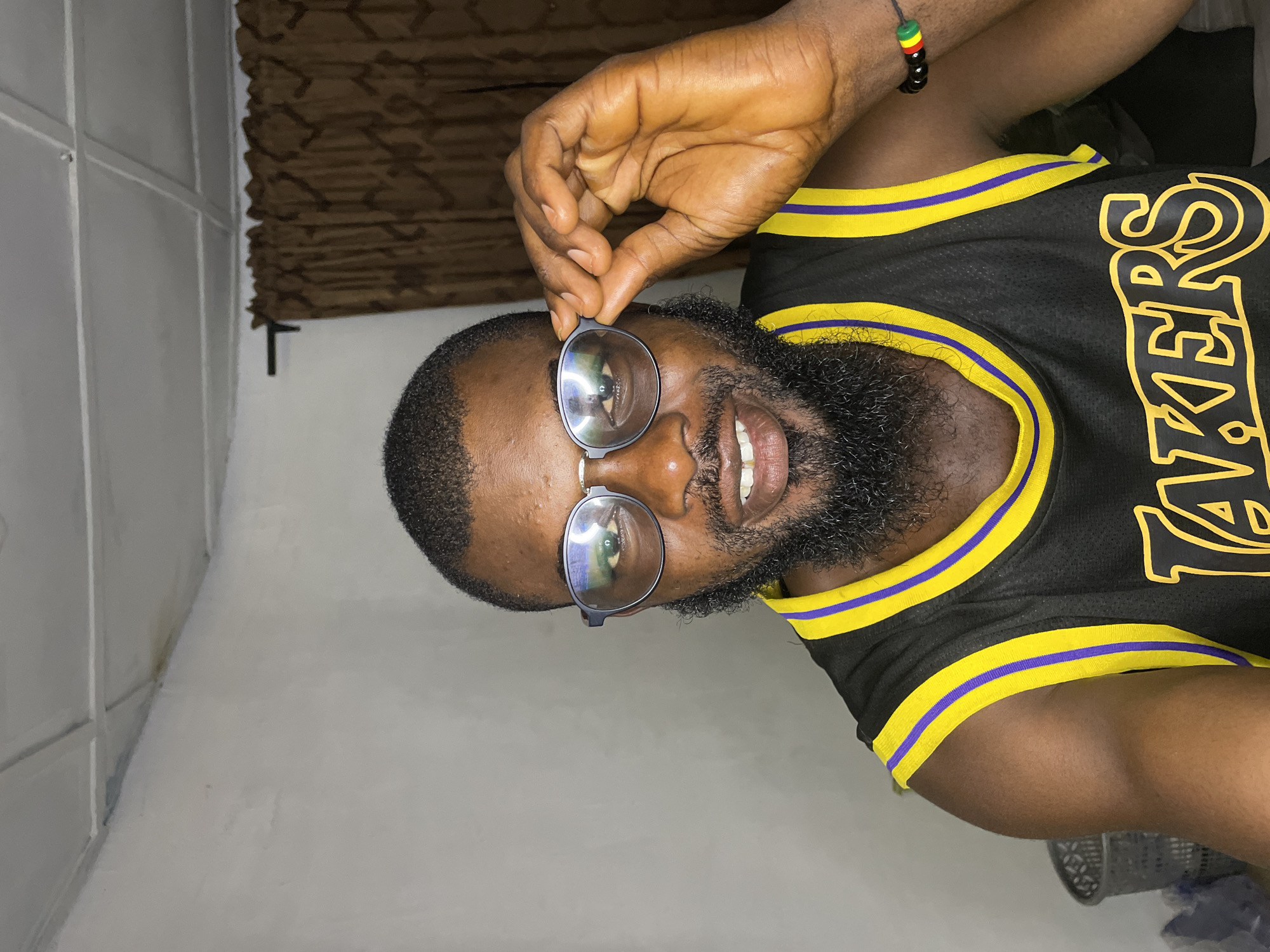 A person is pictured in a close-up shot, the image oriented sideways. The person has dark skin, short dark hair, and a full beard. They are wearing round-framed glasses, and their right hand is holding the glasses up. Their mouth is open in a smile, revealing their teeth. They are wearing a black Los Angeles Lakers jersey with yellow and purple trim. On their left wrist, they have a beaded bracelet with red, yellow, and green beads. The background appears to be an indoor setting with a white wall on the left and a brown textured curtain or blind in the upper left portion of the frame. The lighting suggests it is likely nighttime or indoors with artificial light. There are no external environmental cues visible. The person's expression and posture suggest a casual and perhaps playful mood. The visible text on the jersey reads "LAKERS". The location context is Jos, Nigeria.