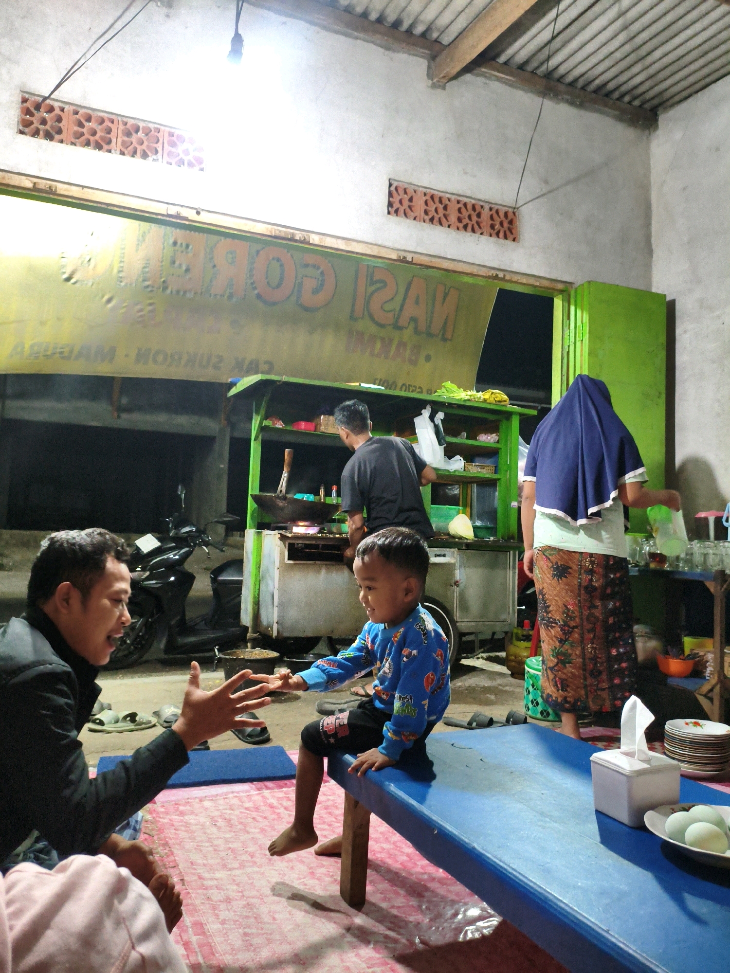 In Krajan Lor, Indonesia, at dusk, a man in a black jacket sits on a pink mat, gesturing with his open palm towards a young child in a blue, long-sleeved shirt with a cartoon print, who is seated on the edge of a blue table. The child's legs are visible, bare from the knee down. Behind the child, a woman wearing a dark blue hijab and a patterned skirt stands near a food stall. The stall, painted green and white, is open for business, with a man in a black t-shirt cooking over a wok. A motorcycle is parked behind the man. The scene is illuminated by overhead lighting. A sign in the background reads "GOR..." in large orange letters on a yellow background.