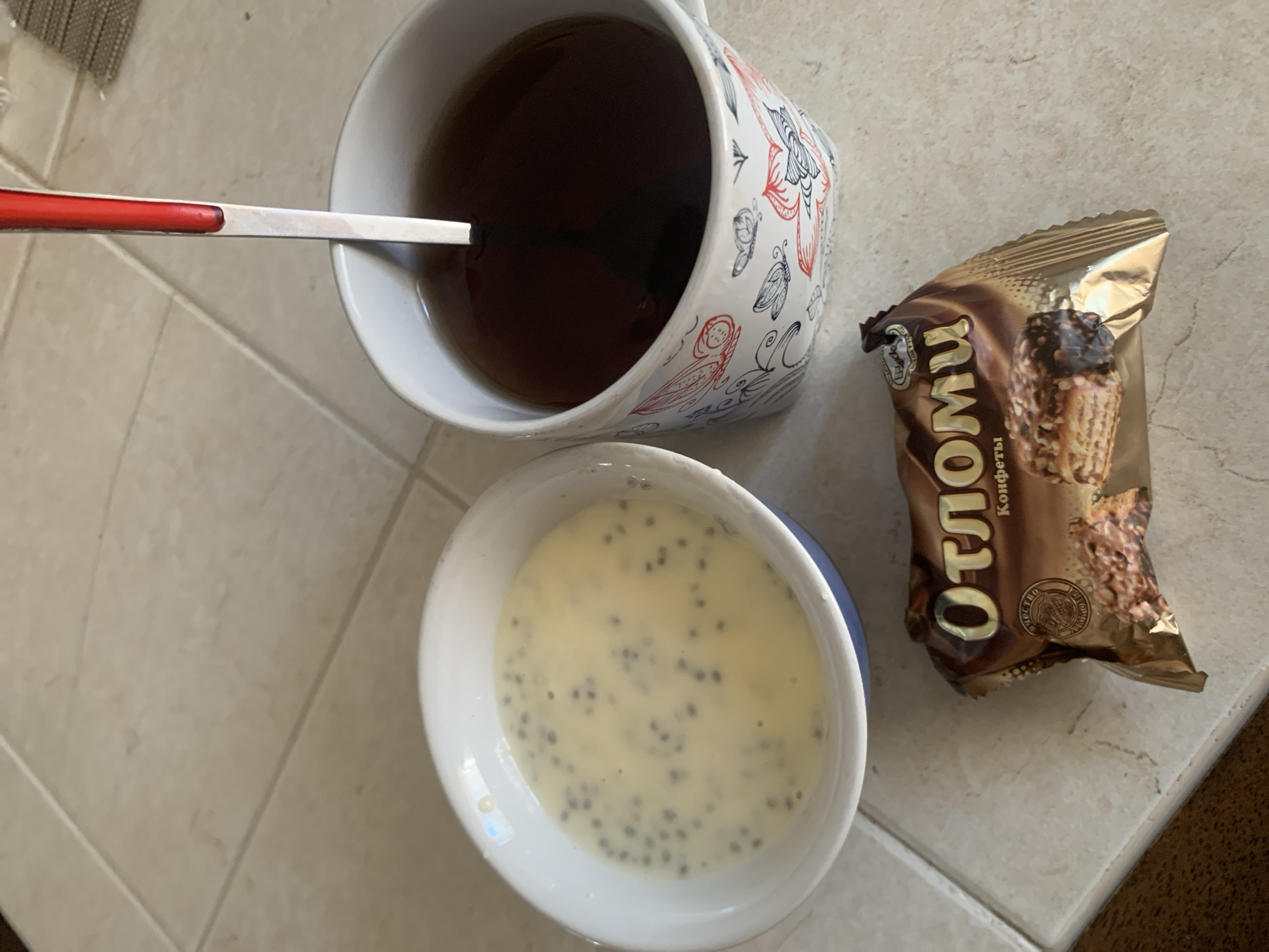 In Kapyĺ, Belarus, on a tiled floor, a breakfast scene is depicted. A white ceramic bowl contains a creamy, pale yellow liquid with dark specks, likely a pudding or yogurt with seeds. To the right, a white mug with a decorative pattern of red and black insects and flowers holds dark tea, with a silver spoon with a red handle partially submerged. In the foreground, a foil-wrapped chocolate wafer cookie with the brand name "Отломи" (Otomi) is visible, its packaging partially opened to reveal the cookie inside. The lighting suggests it is daytime.