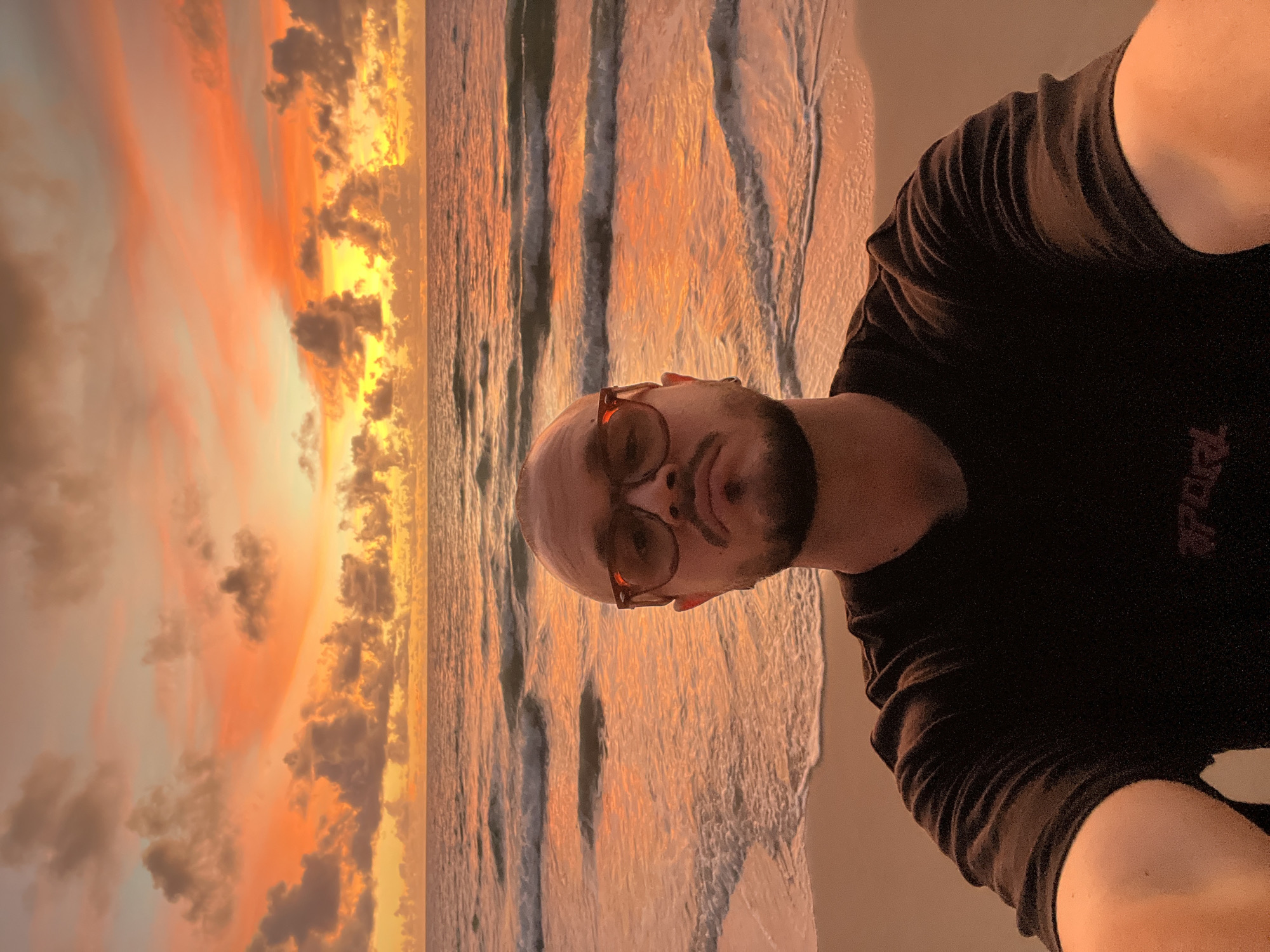 A man with a shaved head and glasses is taking a selfie on a beach at sunset. The sky is filled with vibrant orange and yellow clouds, and the ocean has gentle waves. He is wearing a black t-shirt. The sand is visible in the foreground and the background.