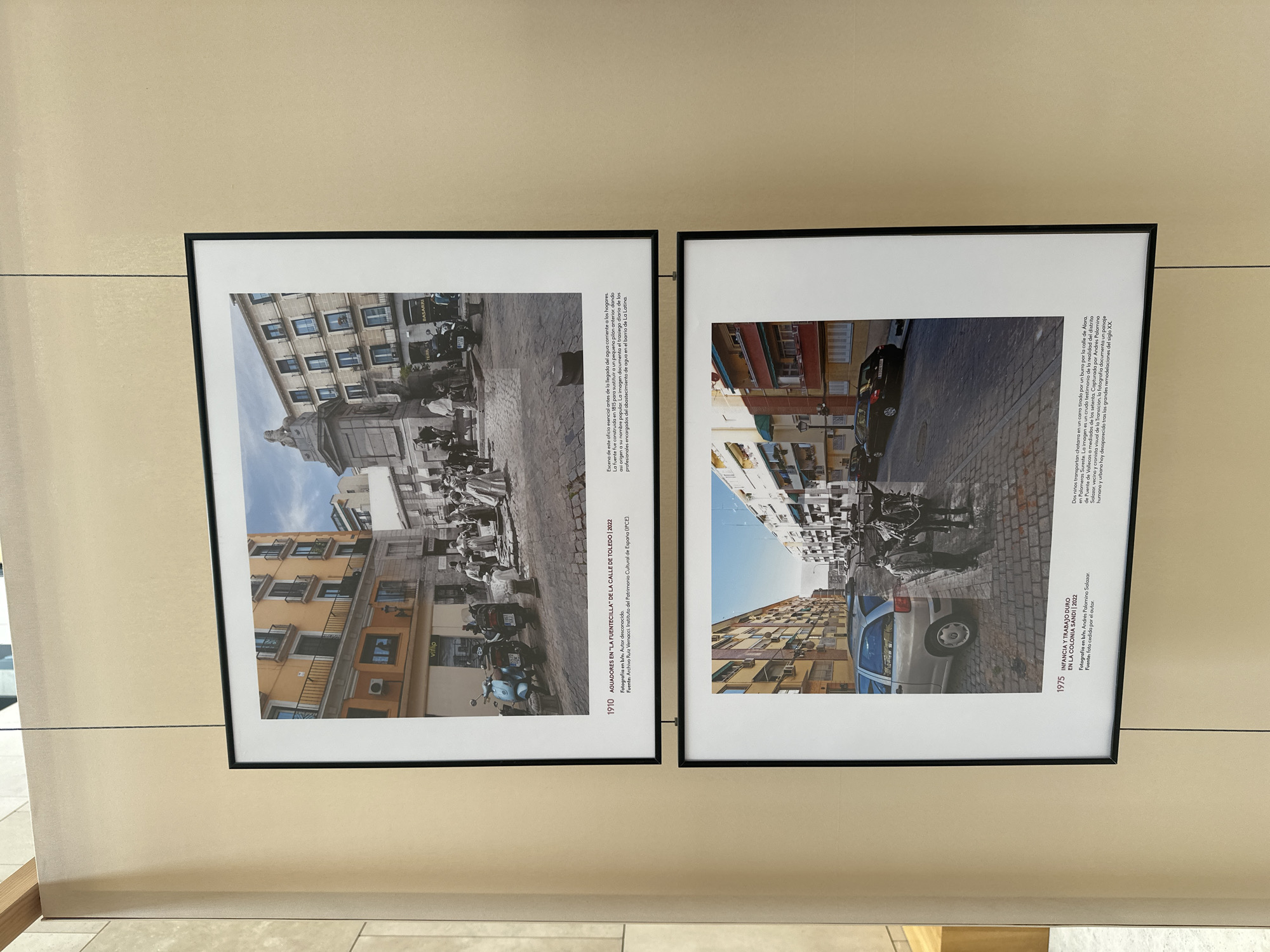 The image displays two framed photographs hanging on a beige wall. The upper photograph, dated 1910, depicts a street scene in Madrid, Spain, possibly near "La Fuencilla" on Calle de Toledo. It shows several people, some in what appear to be historical attire, gathered around a monument. Scooters are parked on the cobblestone street, and buildings with balconies line the background. The text below this photograph identifies it as "AGUADORES EN 'LA FUENTECILLA' DE LA CALLE DE TOLEDO | 2022" and provides details about the photograph's origin and attribution.
The lower photograph, dated 1975, is a black and white image of a street in the Colonia Sandi neighborhood of Madrid. It shows a silver car and a black car parked on a cobblestone street, with a boy and a donkey pulling a cart in the foreground. Buildings with balconies and windows are visible on either side of the street. The text below this photograph describes it as "INFANCIA Y TRABAJO DURO EN LA COLONIA SANDI | 2022" and explains the context of the image, noting children transporting churns by donkey and the documentation of the district's transformation.