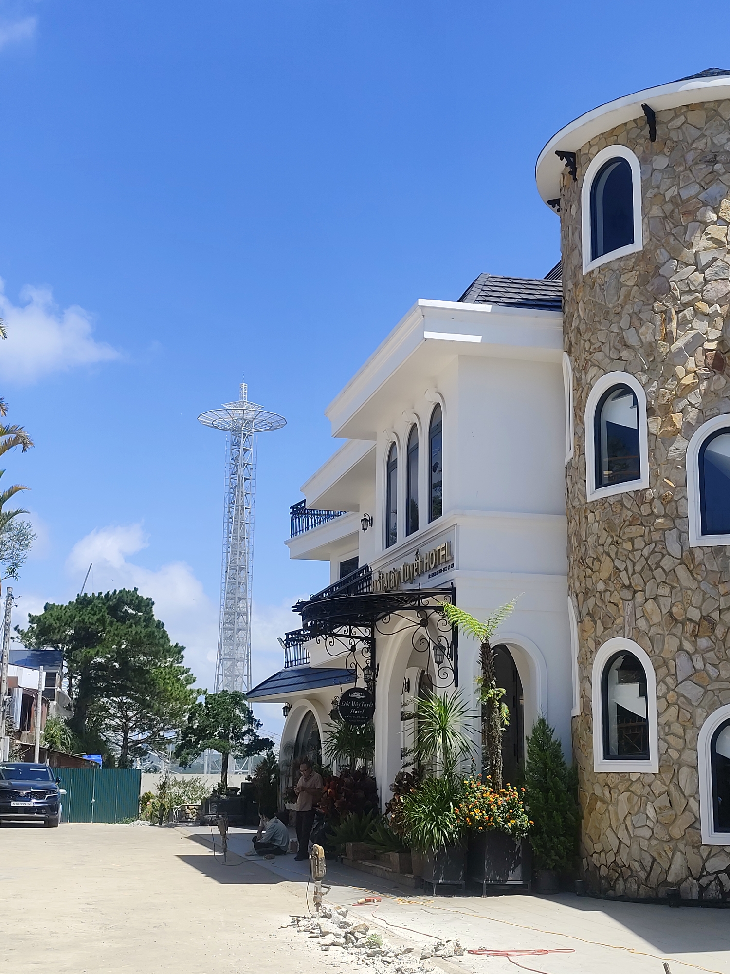 The image depicts a street scene in Da Lat, Vietnam, with a focus on a hotel with a stone-clad tower. A tall, white transmission tower stands prominently in the background against a clear blue sky with sparse clouds. The hotel building features white walls, arched windows with dark frames, and a black tiled roof. The stone tower is round with several arched windows, also with dark frames. The hotel entrance has a black wrought-iron awning and a sign that reads "Dai May Tuyet HOTEL". In the foreground, there is a dirt road with some construction debris and what appear to be construction tools, including jackhammers. Two men are visible near the construction area, one sitting and the other standing. A black SUV is parked on the left side of the road. Lush green trees and palm trees are scattered throughout the scene, contributing to a tropical atmosphere.