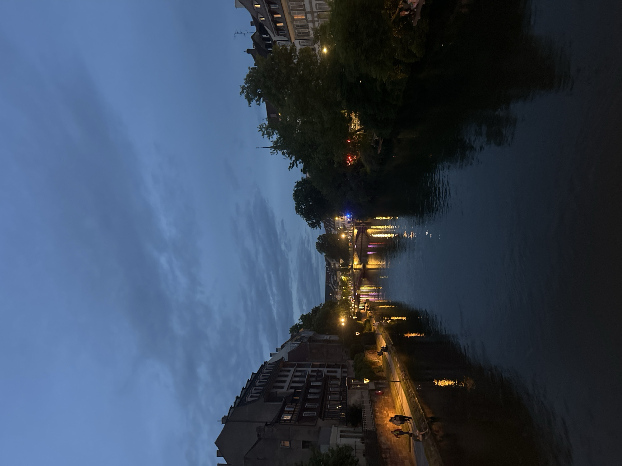 This nocturnal image, taken in Strasbourg, France, depicts a serene evening scene along a river. The overall mood is dim, with the sky showing shades of dark blue and grey, indicating either dusk or early night, accompanied by visible clouds.
The river, a prominent feature, flows from the foreground towards the background, reflecting the numerous artificial lights from the buildings and streetlamps lining its banks. On the left side of the river, a row of multi-story buildings with traditional European architecture extends along the waterfront. Some windows are illuminated, suggesting activity inside. A paved walkway runs alongside these buildings, where several people can be seen as dark silhouettes, strolling or standing. Streetlights illuminate parts of this path and the water directly below.
On the opposite bank, dense trees obscure parts of the buildings behind them, though some structures, including those with illuminated windows, are visible through and above the foliage. Further down the river, multiple bridges span the water, adorned with lights that cast colorful reflections onto the rippling surface. The lights from buildings and bridges create streaks and patterns of yellow, orange, and hints of red and blue in the dark water. The water itself appears calm but with small ripples, capturing and distorting the light reflections.
The scene is peaceful and atmospheric, showcasing the city's charm under the evening sky. There is no visible text in the image.