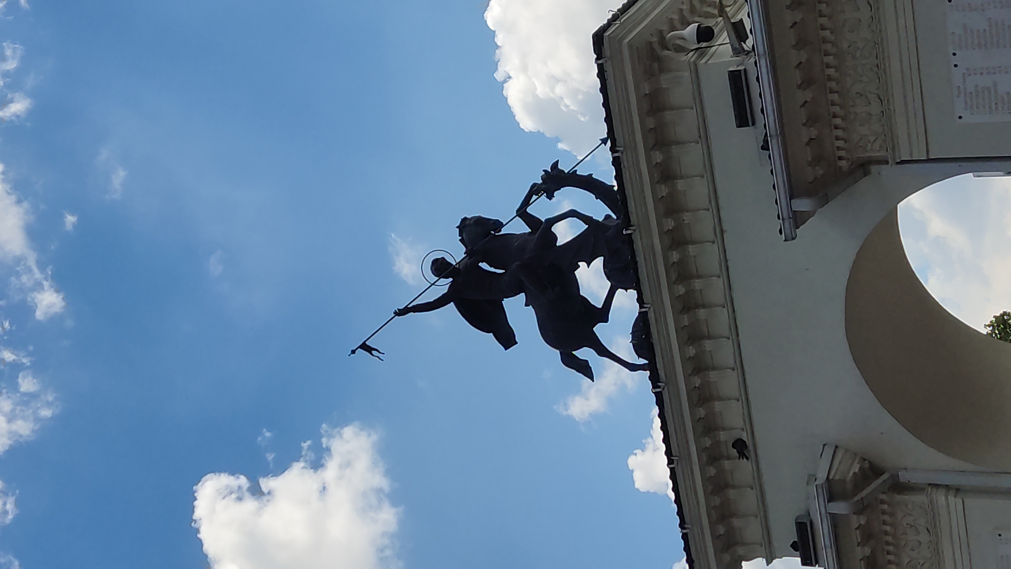 This media file features a silhouette of a statue against a bright blue sky with scattered white clouds. The statue depicts a figure on horseback, appearing to be in the act of spearing a dragon. The figure has a halo around its head, suggesting a religious or mythological subject, likely St. George. The statue is mounted on an ornate architectural structure that includes columns and decorative molding. The architecture is a light color, contrasting with the dark silhouette of the statue. Part of a large archway is visible on the right side of the frame. The scene is brightly lit, indicating it is daytime, likely midday due to the intensity of the light and the clear sky. The perspective is from below, looking up at the statue and the sky. The overall mood is majestic and heroic, amplified by the upward-looking angle and the dramatic silhouette. This is a public monument, possibly an architectural element of a significant building or a standalone memorial. The crops show details of the statue and the surrounding architecture, confirming the presence of the horse, rider, spear, and dragon. The location context is Krasnodar, Russia, which might hint at the specific historical or cultural significance of this monument within that region. There is no visible text in the image.