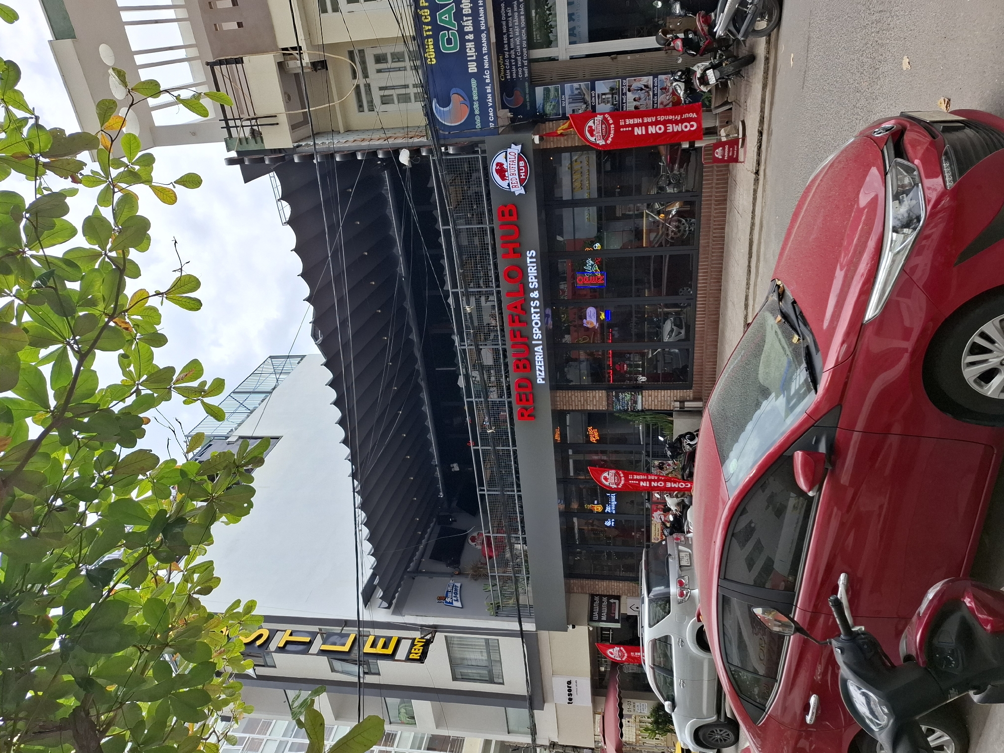 This is a medium shot, eye-level, outdoor, daytime photograph taken on a street in Nha Trang, Vietnam. The scene depicts a multi-story building with awnings extending over a storefront. Dominating the lower half of the frame is a red sedan, parked on the street. To the left of the sedan, a black motorcycle is partially visible.
The building features a prominent red sign with white lettering that reads "RED BUFFALO HUB" and below it, "PIZZERIA | SPORTS & SPIRITS". Further up the building, illuminated yellow letters spell out "STLE" on a white facade, with the word "RENT" below it. The storefront windows show glimpses of the interior, including people, seating, and decorative elements. Several red flags with white text are planted on the sidewalk in front of the establishment, with one reading "COME ON IN... Your friends are HERE!!". A smaller sign near the entrance indicates "PARKING" and "Thank you!".
To the right of the "RED BUFFALO HUB" establishment, another building displays a blue sign with white lettering. The street is paved, and a sidewalk runs alongside the building. Green foliage from overhead trees drapes into the top of the frame. The sky appears overcast. Parked motorcycles are visible further down the street to the right, and a silver SUV is parked in front of the "RED BUFFALO HUB". The overall impression is a lively street scene in a commercial area.