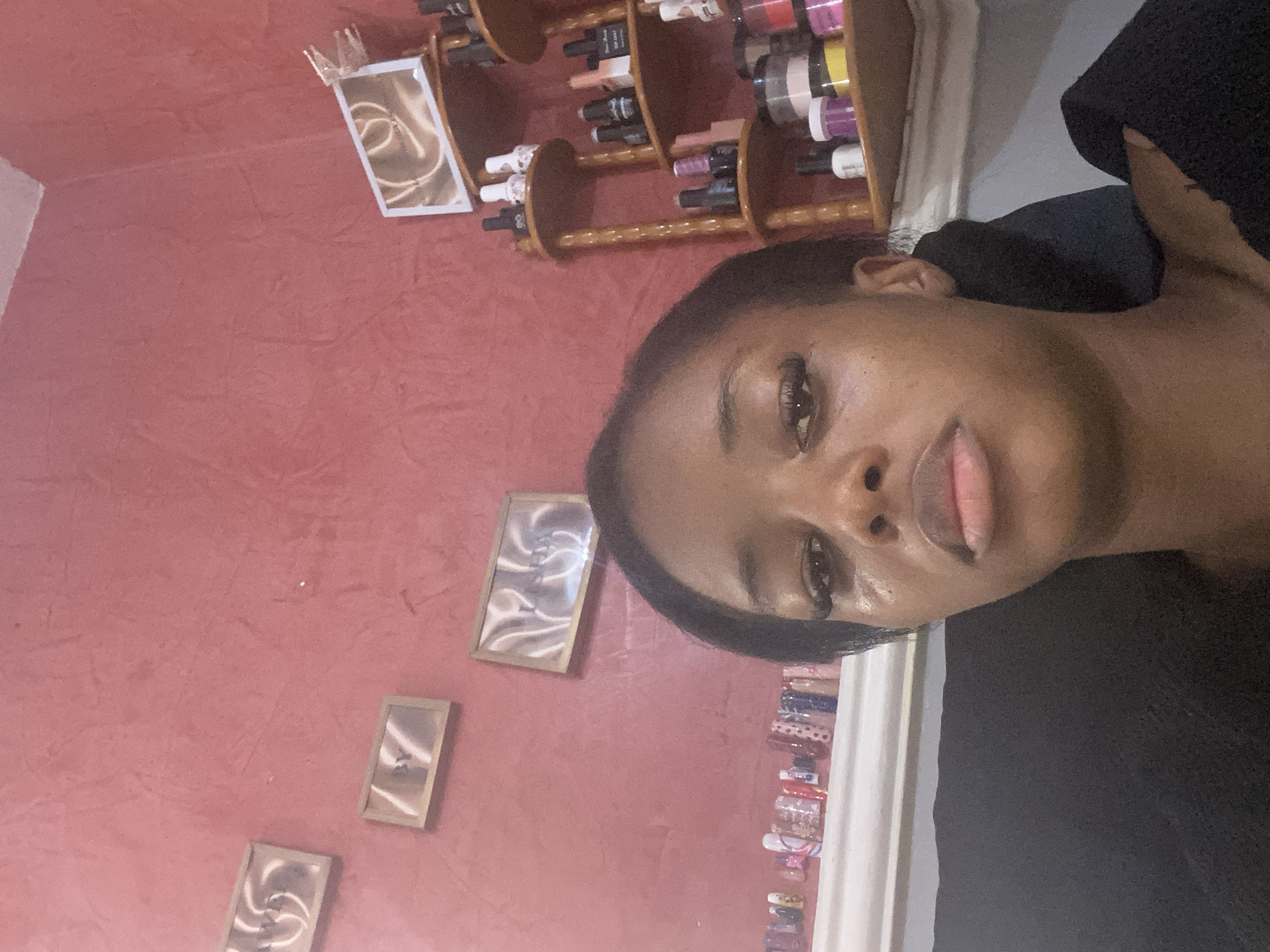 The image depicts a close-up of a young Black woman's face, positioned in the foreground. She has dark skin, dark hair styled back from her face, and is wearing makeup, including false eyelashes. Her expression appears neutral to slightly solemn.
In the background, the wall is painted a textured reddish-pink color. To the left of the woman, there are three framed pictures hanging on the wall. The topmost frame contains an abstract design. Below it, the second frame displays the letters "BY" in a stylized font. The third frame, positioned further to the right, also contains an abstract design.
Below the framed pictures, a shelf runs horizontally across the wall. This shelf is lined with a collection of artificial fingernails, displayed in various colors and designs.
To the right of the woman, a wooden shelving unit with multiple tiers is visible. This unit is filled with various bottles, jars, and containers, likely nail polish, gels, and other beauty products, suggesting a beauty salon or a personal nail art setup. Some bottles are black, white, pink, and yellow. A mirror is also visible on the top shelf of this unit.
The overall setting appears to be an indoor space, possibly a room within a home or a small business in the city of Jos, Nigeria. The lighting is somewhat dim, creating soft shadows. The dark fabric in the lower right corner suggests the woman might be wearing a dark top or is positioned against a dark background.