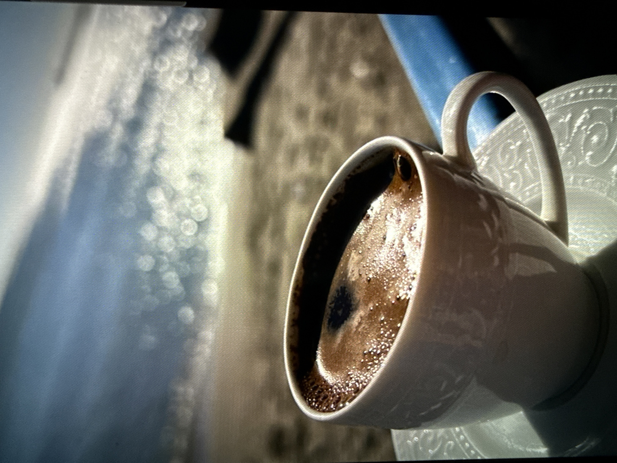 A close-up photograph shows a white ceramic coffee cup filled with dark coffee on a matching saucer. The cup is tilted slightly towards the viewer. The saucer has a raised, embossed pattern of swirls and circles. Light reflects off the surface of the coffee. The background is out of focus, showing a blurred surface and a blue-toned object or area. The location and city are unknown.