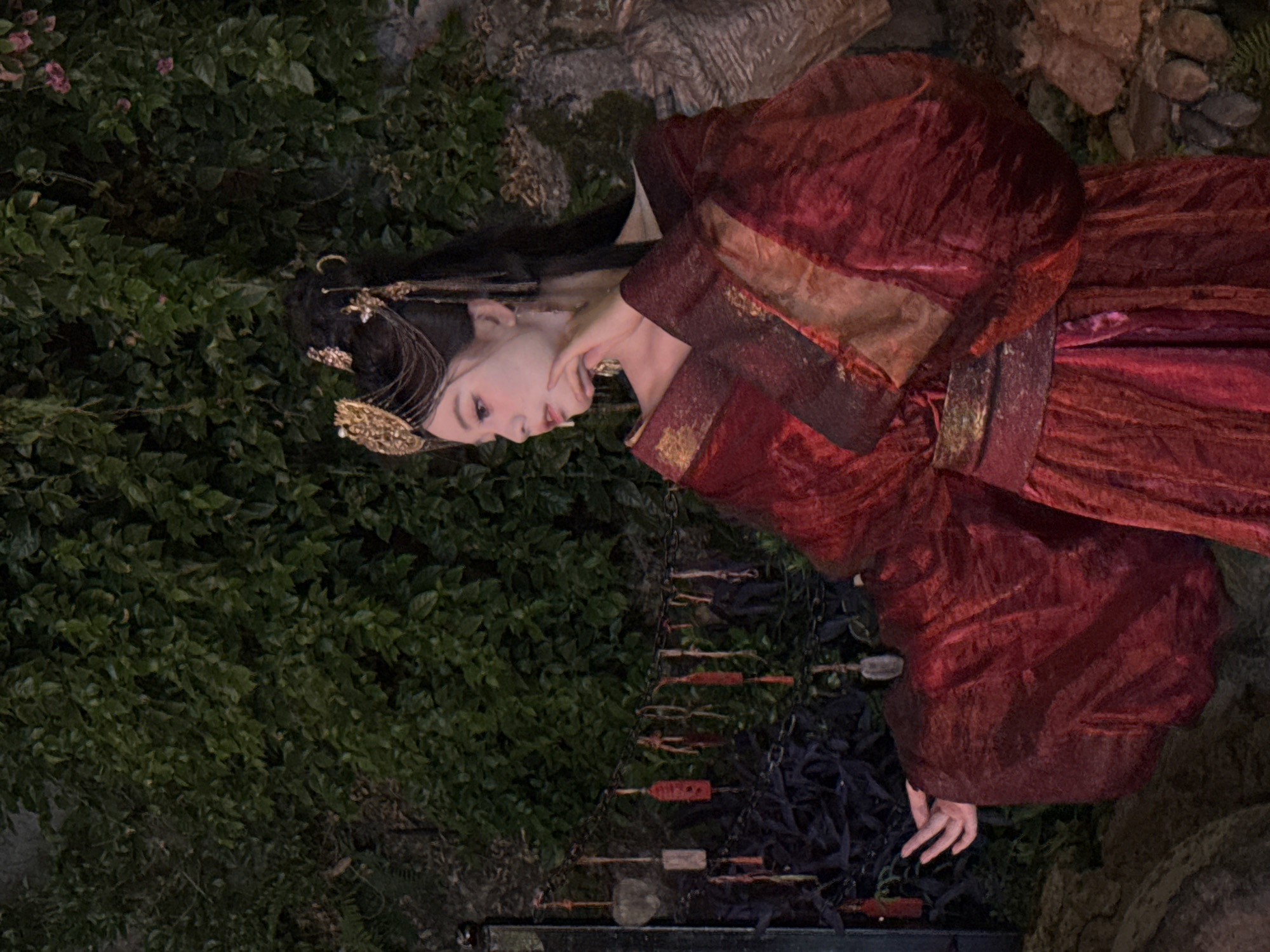 A young woman in traditional Chinese attire stands against a backdrop of lush green foliage and stone. Her red and gold robe features wide sleeves and a sash, with ornate gold hairpins and chains adorning her dark hair. She gazes to her left, her hand near her face. A chain with small red and white tags hangs to her left, and various plants and rocks fill the foreground. The scene appears to be an outdoor setting, possibly a garden or historical site, within Yuzhong District, China.