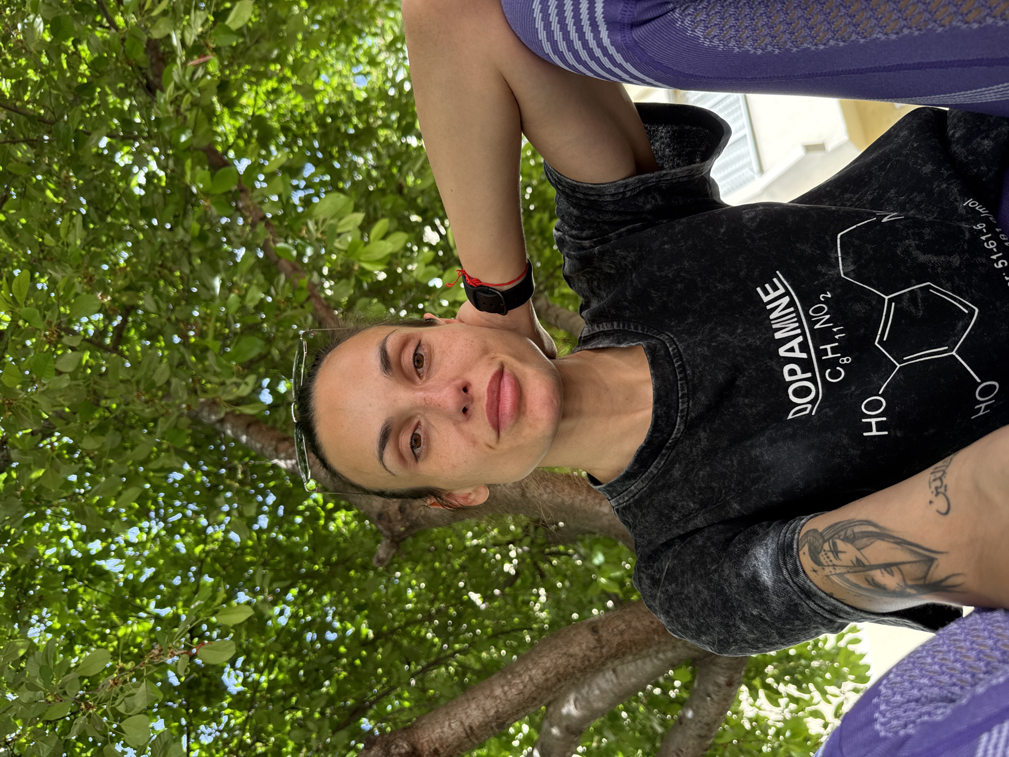 A woman wearing a black t-shirt with a dopamine chemical structure design and purple patterned leggings is standing in Pleven, Bulgaria. The camera is angled up towards her, capturing the green foliage of a tree behind her. She has a black watch and a red bracelet on her left wrist, and a tattoo on her left forearm. Sunglasses are perched on top of her head.