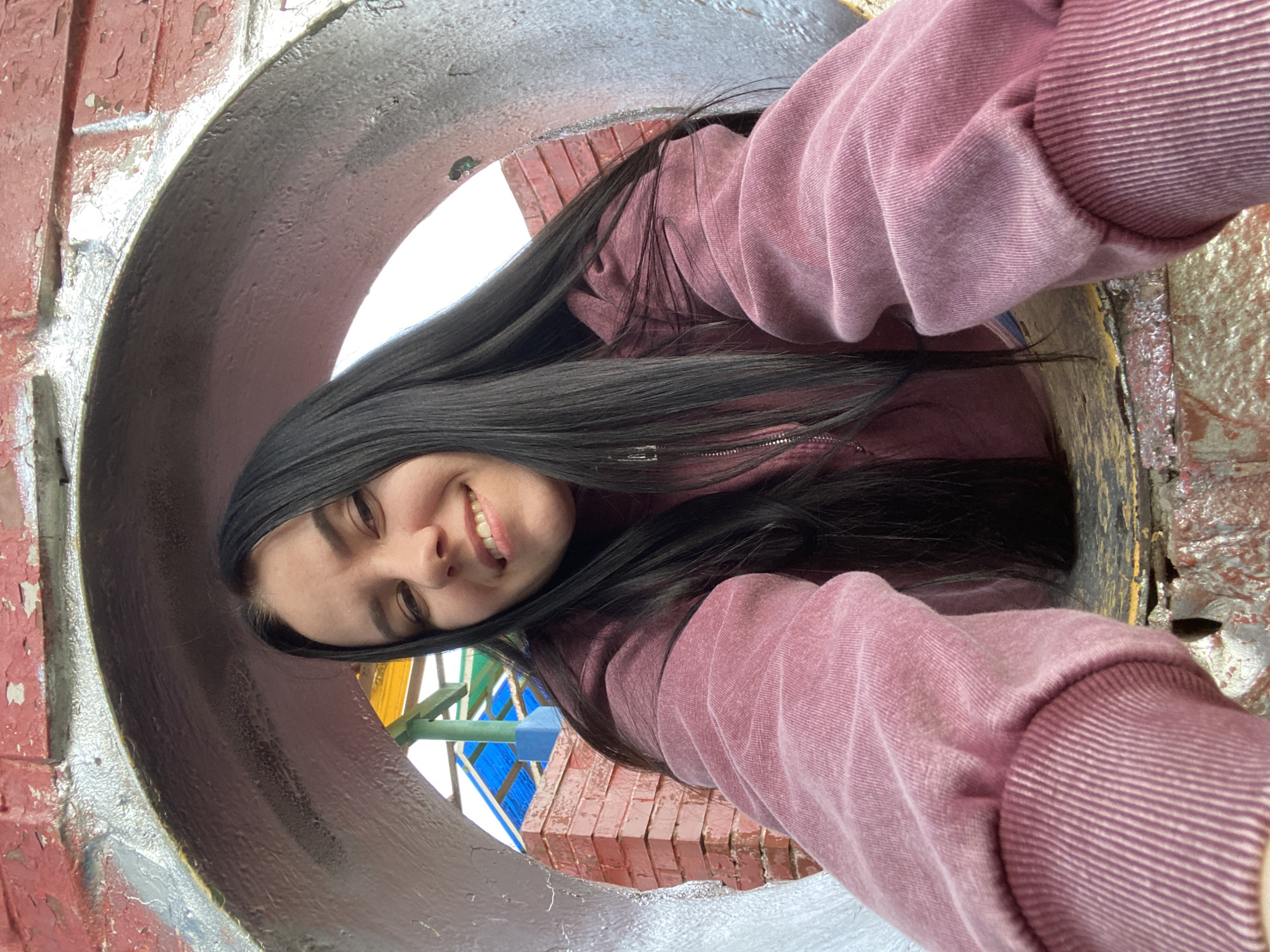 A person with long, dark hair and wearing a pink sweatshirt is looking through a large circular opening, likely a pipe or tunnel. The opening is made of a material that appears to be painted metal or concrete, with visible red brick on the surrounding surface. The person is smiling at the camera, suggesting they are taking a selfie. In the background, visible through the opening, there are structures with colorful geometric patterns, including yellow, green, and blue. This scene could be in Perm, Russia, as the geometric structures are characteristic of some playground and recreational installations there. The person is stationary, and the action is centered on capturing the photo.