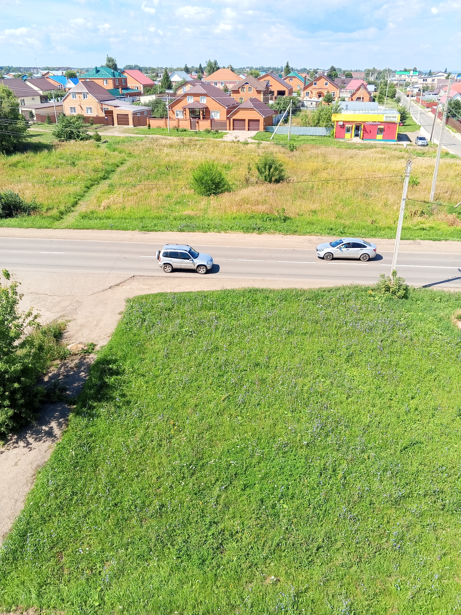 An aerial shot captures a suburban landscape under a partly cloudy sky. A two-lane asphalt road traverses the frame horizontally. On the road, a silver SUV is visible in the left lane, and a silver sedan is in the right lane, both moving away from the viewer. The road is bordered by a grassy verge, transitioning into a field of dry grass and small bushes on the left. On the right side of the road, a red and yellow building with a yellow roof and signage stands, possibly a small store, with a single car parked in front of it. Beyond the building and the field, a residential area with numerous houses is spread out, featuring various roof colors and architectural styles. The houses are surrounded by green lawns and trees. The overall scene depicts a sunny day in a developed, yet somewhat rural, area.