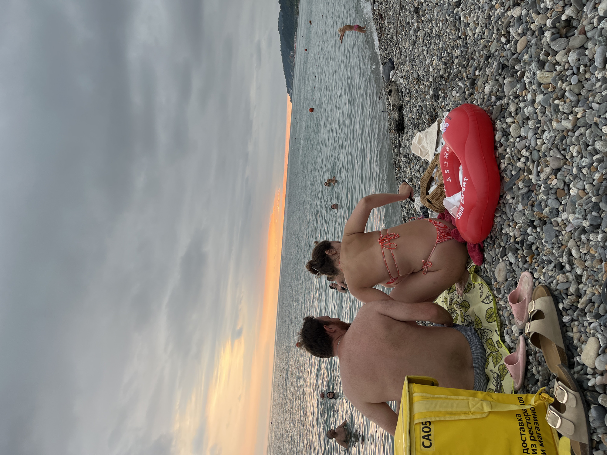 This image depicts a coastal scene in Sochi, Russia, likely during the late afternoon or early evening, indicated by the soft, warm glow of the sunset on the horizon. The foreground is occupied by a couple sitting on a patterned beach towel on a pebble-strewn shore. The man, facing away from the camera, has his back bare and is wearing shorts. The woman, also facing away, is in a red and white patterned bikini. Beside them is a bright yellow cooler bag with the label "CAO5" and Cyrillic text. Scattered on the pebbles are several pairs of sandals and a red inflatable swim ring.
In the background, the sea is calm, with several people visible swimming or wading. The water is a deep blue-green, reflecting the overcast sky, which is a mix of grey clouds with hints of orange and yellow near the horizon. A distant coastline with a verdant, tree-covered hill is visible on the right side of the frame. A few red buoys dot the water. The overall atmosphere suggests a relaxed recreational setting by the Black Sea.